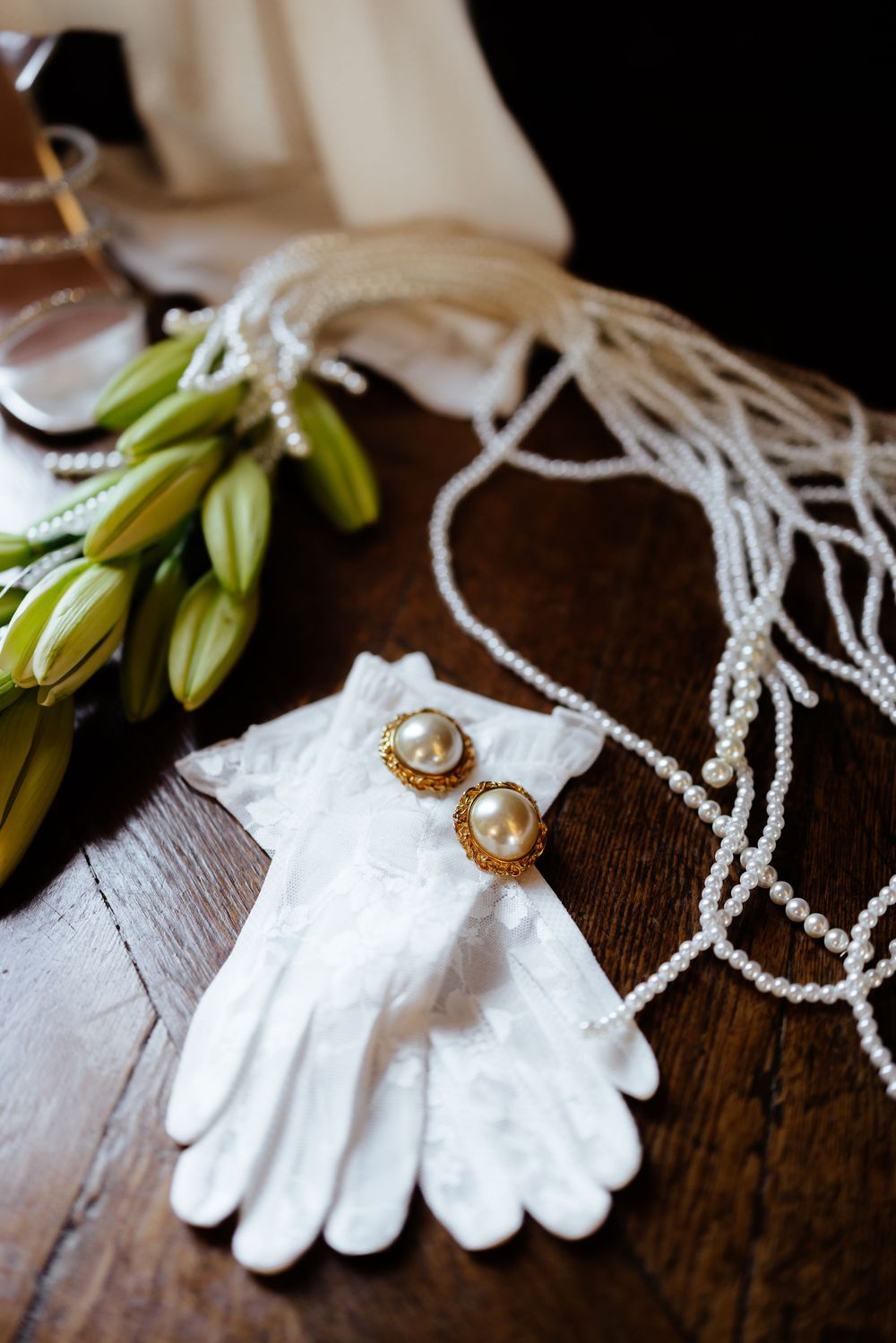 White gloves and pearl earrings displayed with green lilies and beaded necklaces on a wooden surface.
