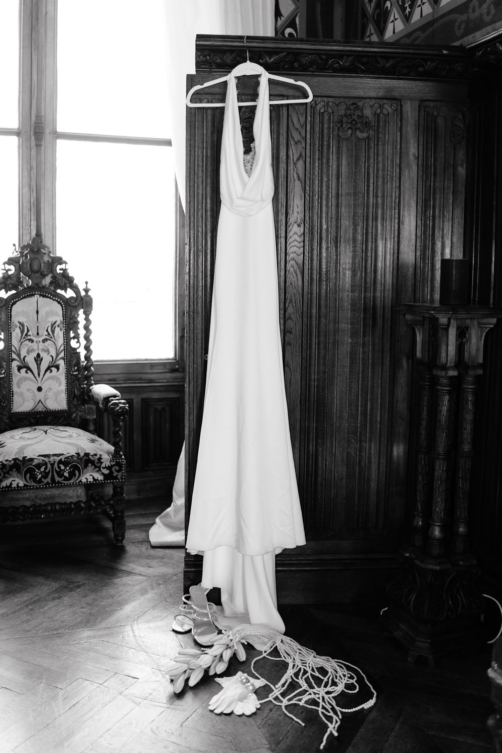 A white wedding dress hangs elegantly against dark wood paneling with vintage-style shoes placed beneath it.