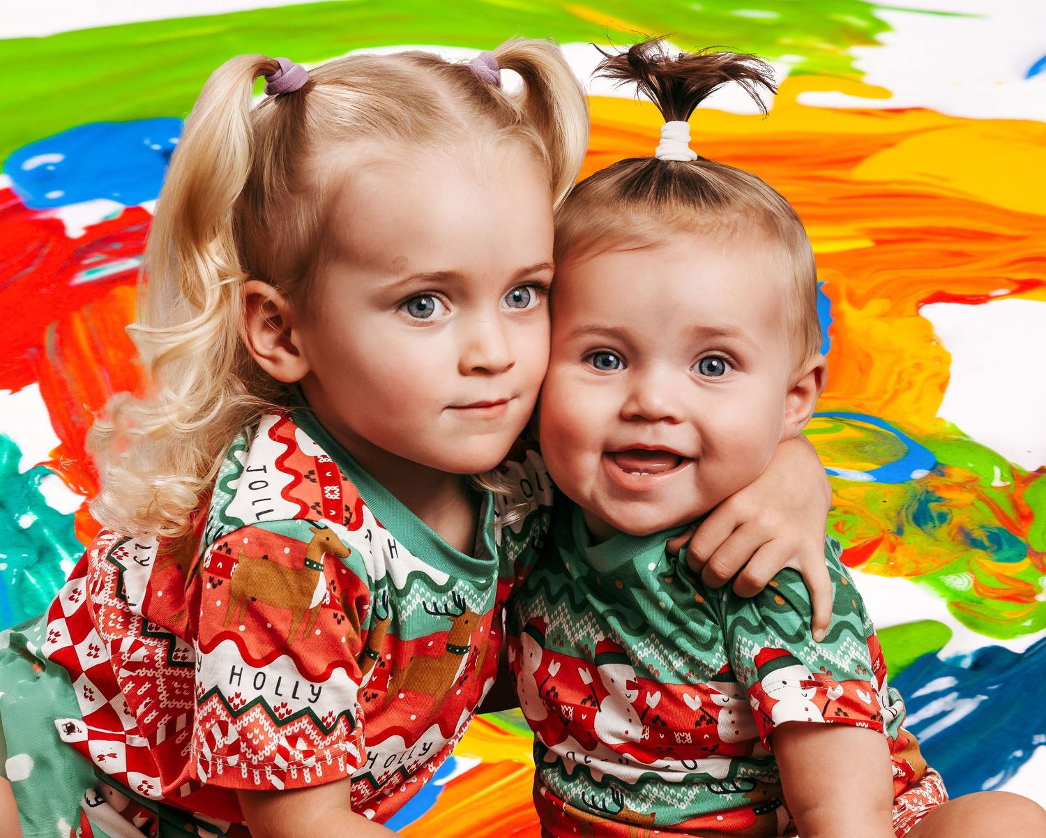 Two young children wearing festive holiday outfits pose together against a colorful painted background.
