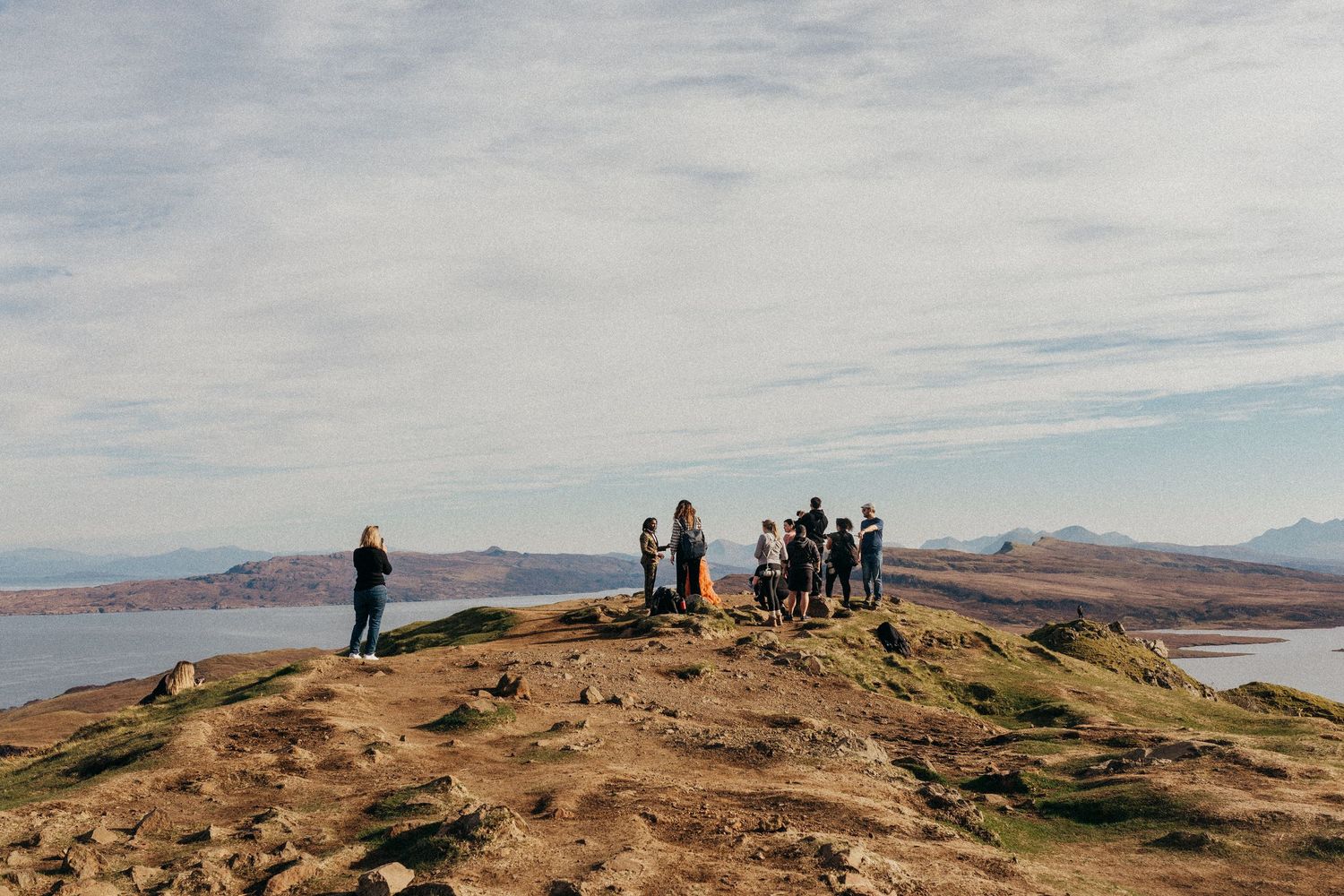 Cloudy skies over a dried grassy coastal hill with people hiking above a serene bay in the distance.