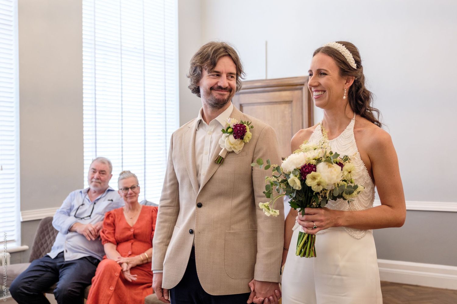 A happy wedding couple shares a joyful moment together while holding a bouquet in a bright indoor venue.