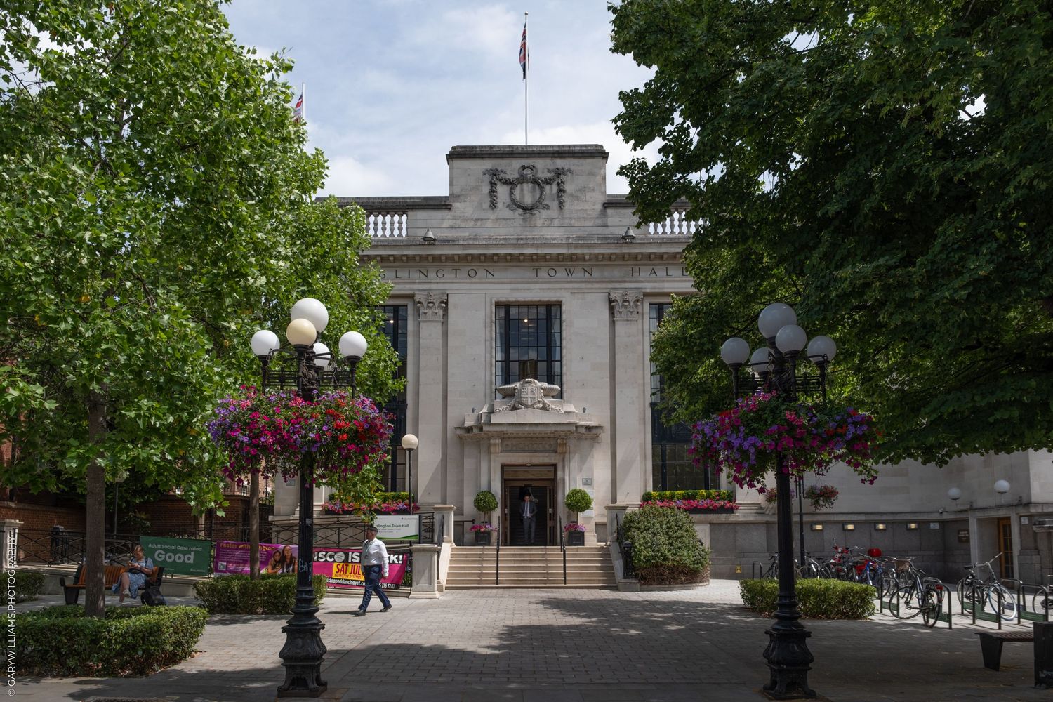 Grand stone entrance to a government building with decorative light posts and hanging flower baskets along the pathway.