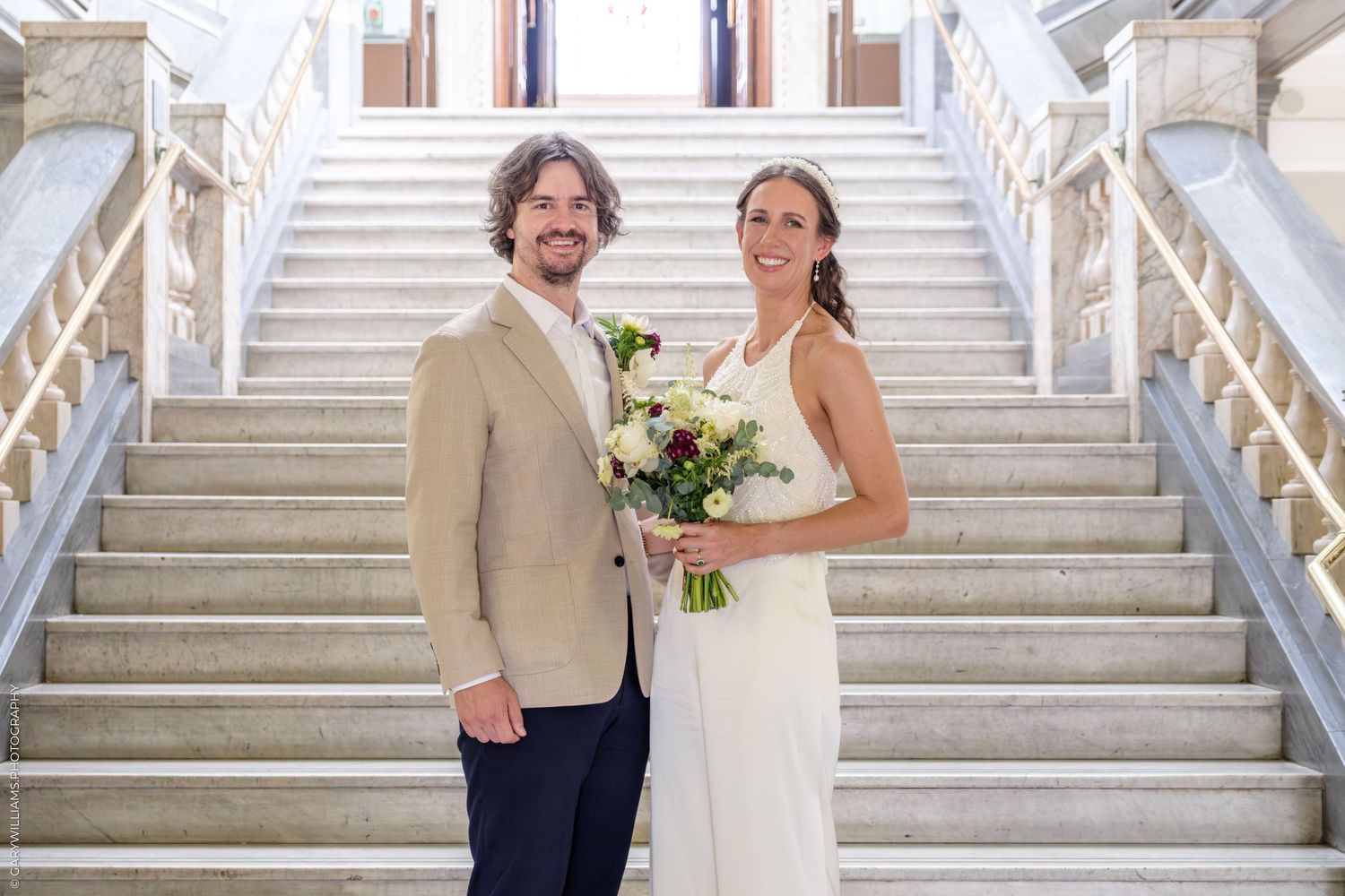 A couple poses for a wedding photo on grand marble stairs with white railings and a traditional venue backdrop.