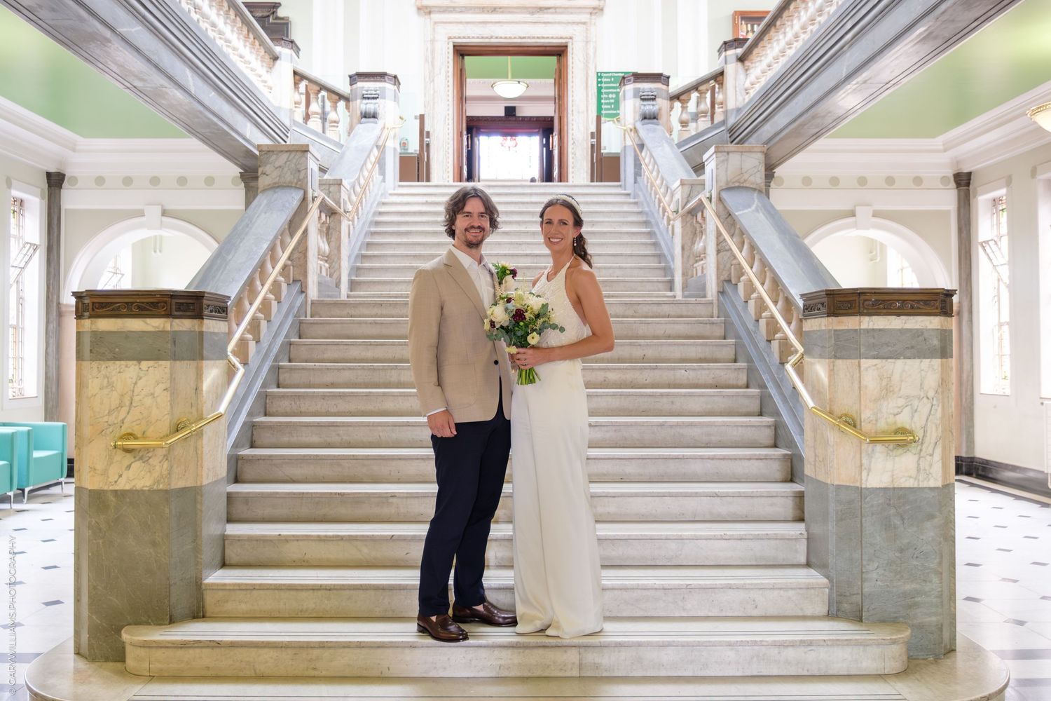 A couple stands together on an elegant marble staircase with white railings in a formal interior setting.