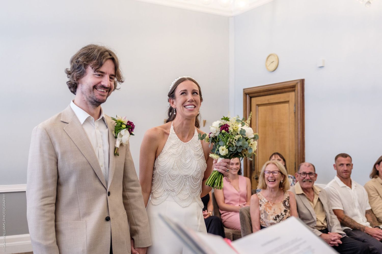 A joyful newlywed couple stands together during their wedding ceremony while guests look on from the background.