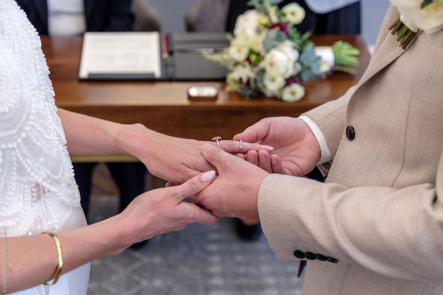 Close-up shot of hands exchanging wedding rings during a ceremony with white dress and tan suit visible.