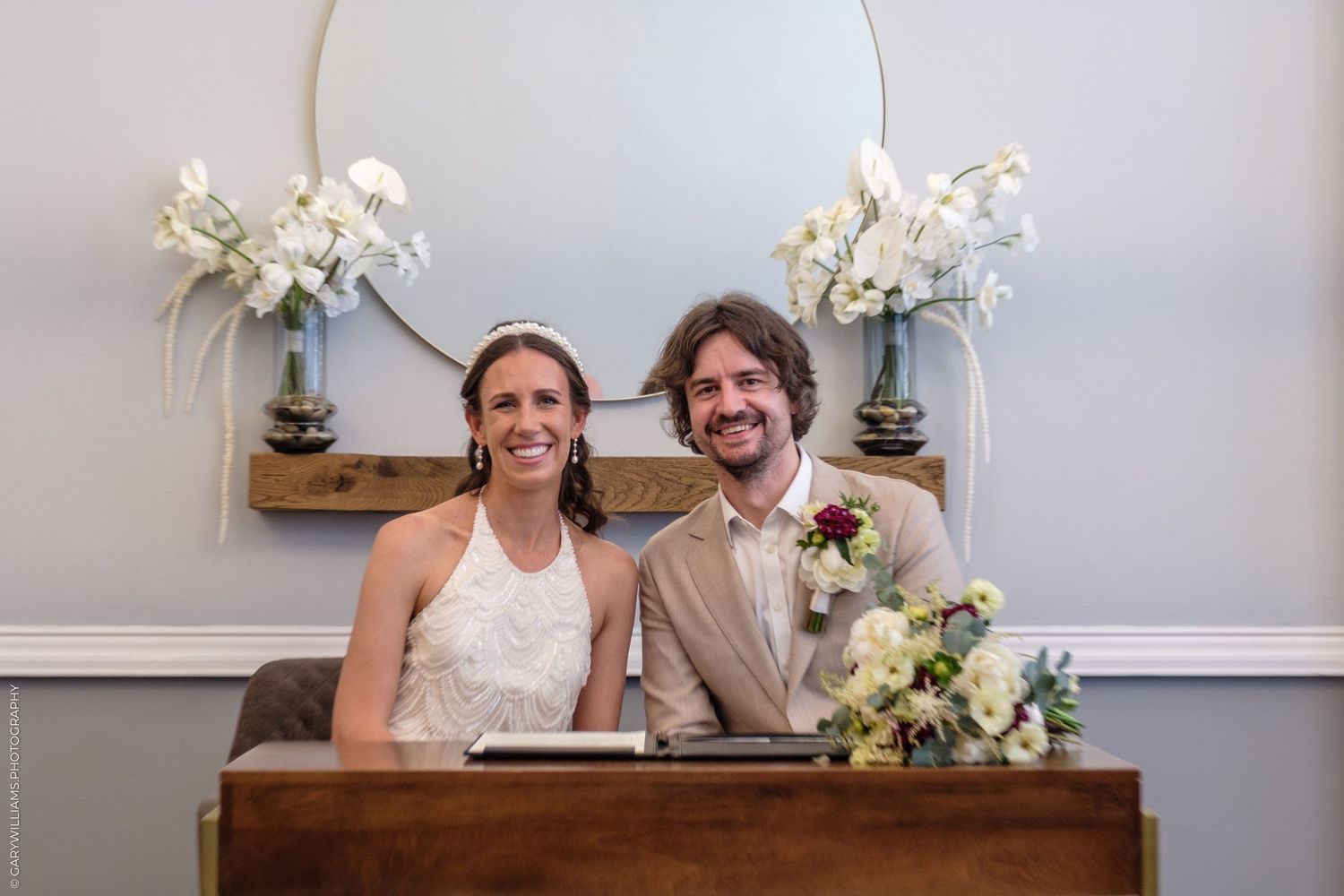 A smiling couple sits at a wooden table with elegant white floral arrangements displayed on either side.