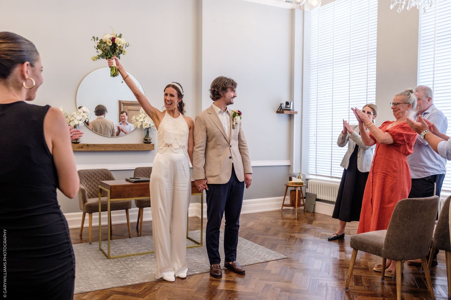 A couple celebrates in a bright indoor venue as guests applaud during an intimate wedding ceremony.