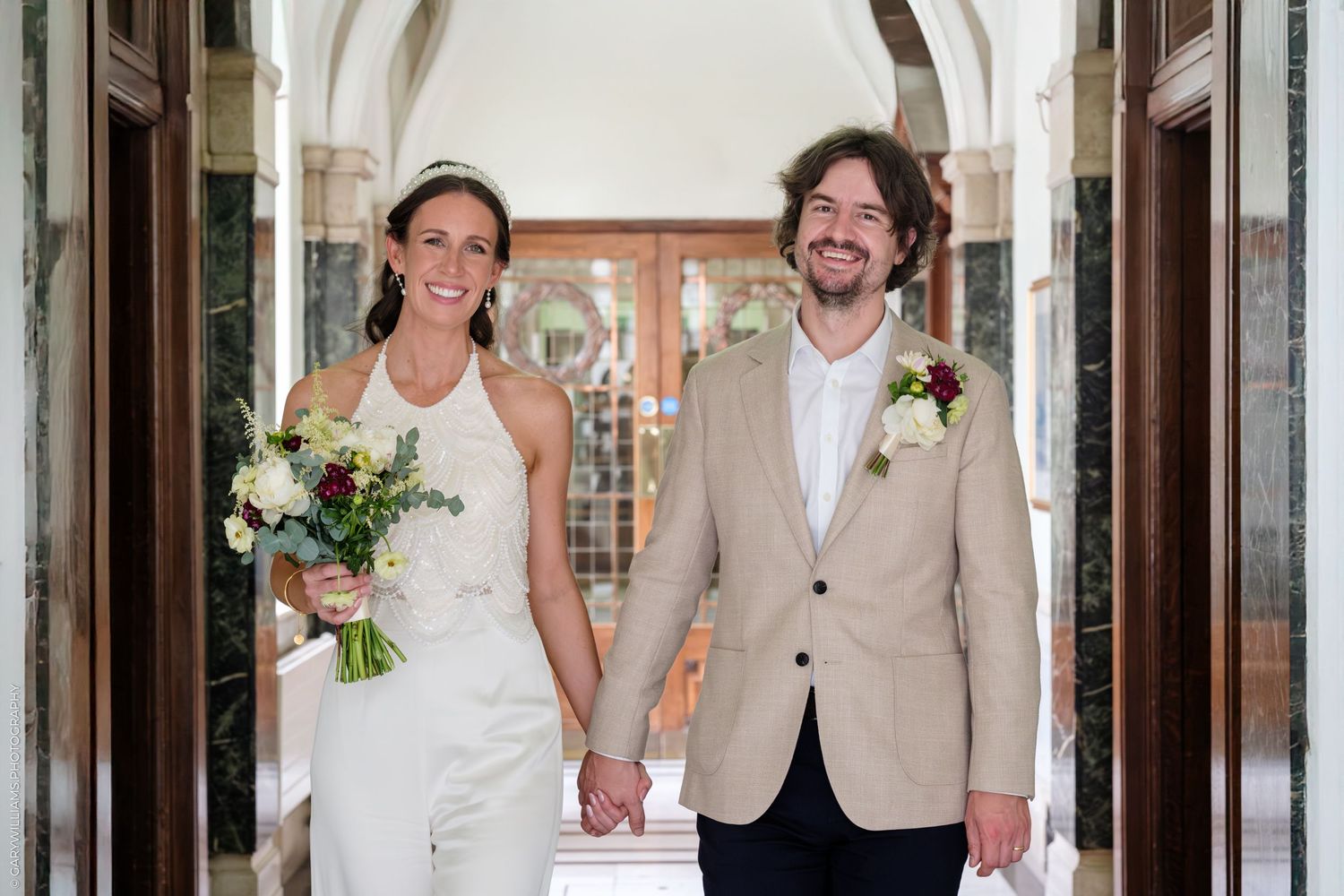 A couple in wedding attire holds hands while walking through an ornate hallway with arches and marble columns.