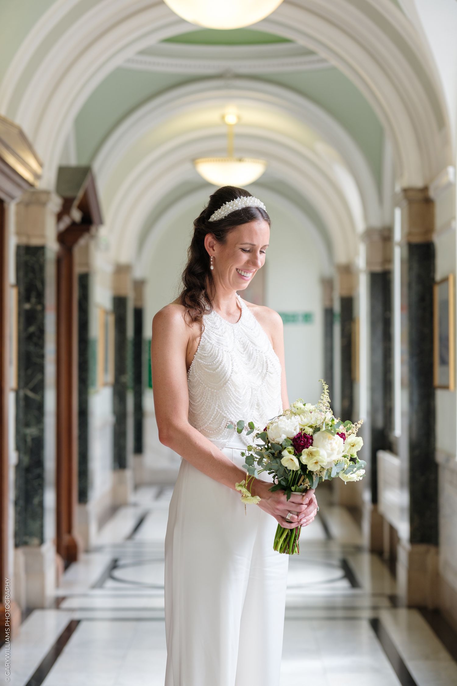 A bride in a white dress smiles while holding a bouquet in an elegant arched corridor with classical architecture.