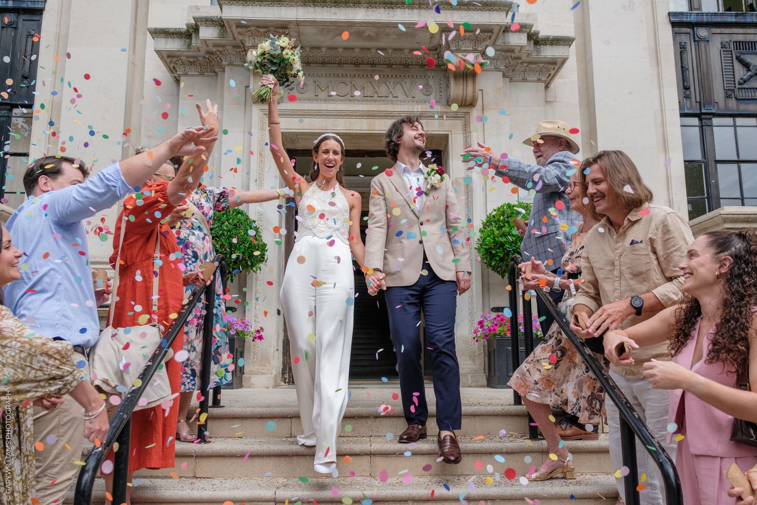 Newlyweds walking down steps while guests throw confetti during outdoor summer wedding celebration.