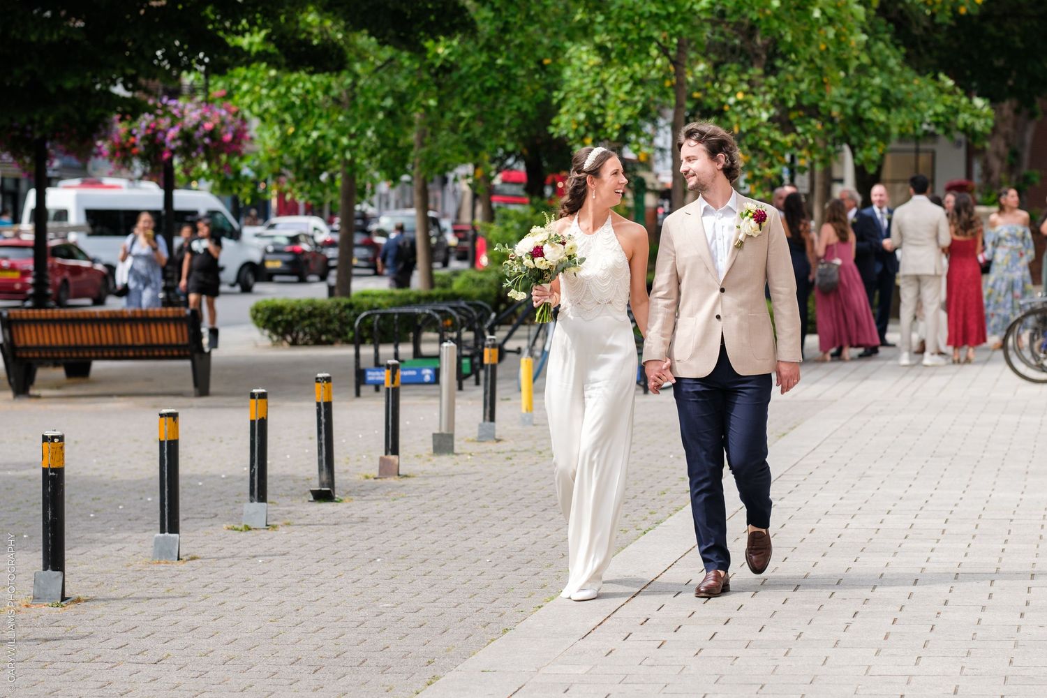 Well-dressed guests stroll along a paved walkway lined with flower planters and decorative bollards at an outdoor event.