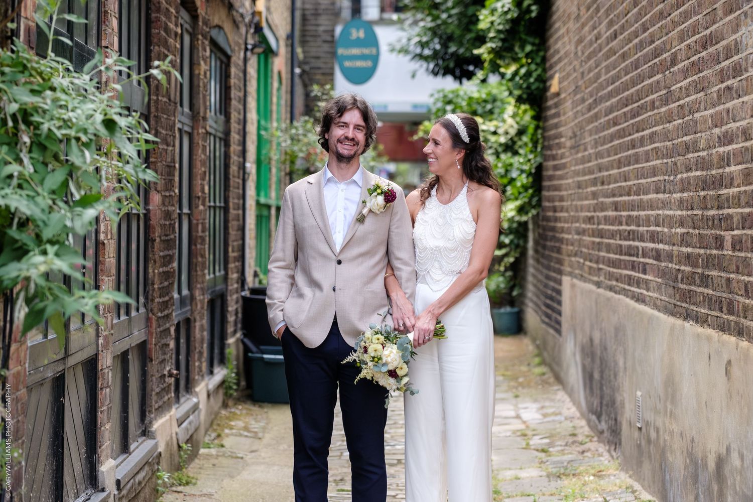 A bride and groom walking together and smiling in a charming brick-lined alleyway with greenery.