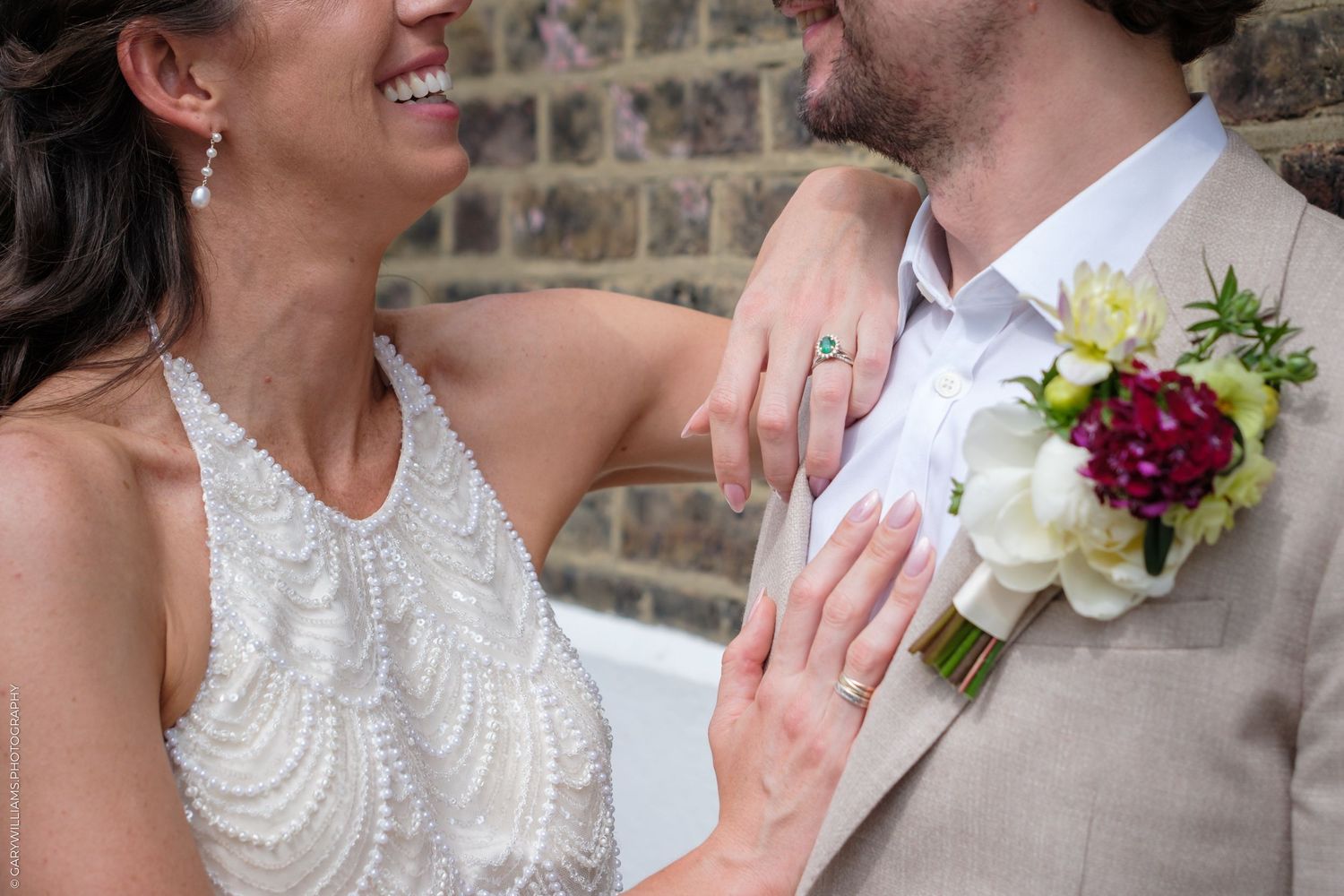 Wedding party members share joyful reactions and embraces while holding floral bouquets against a brick wall backdrop.