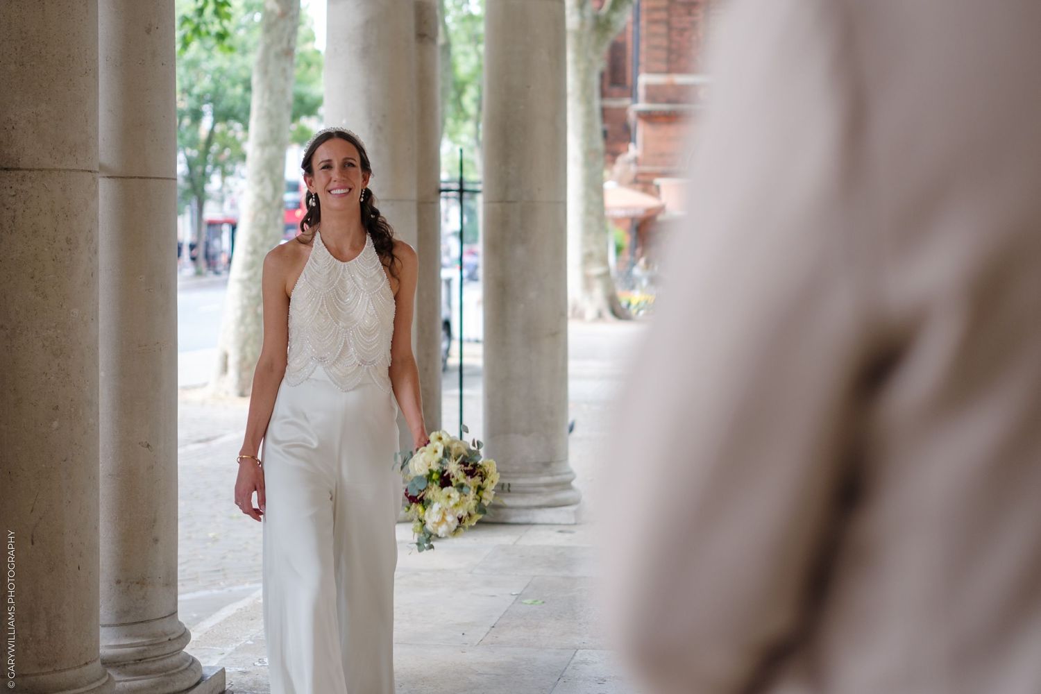 A bride in a white sleeveless wedding dress walks through an elegant columned walkway with a bouquet.