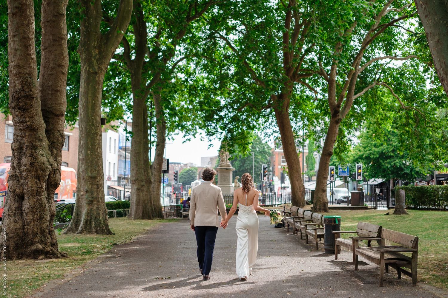 A tree-lined path in a park with benches and people walking on a sunny summer day.