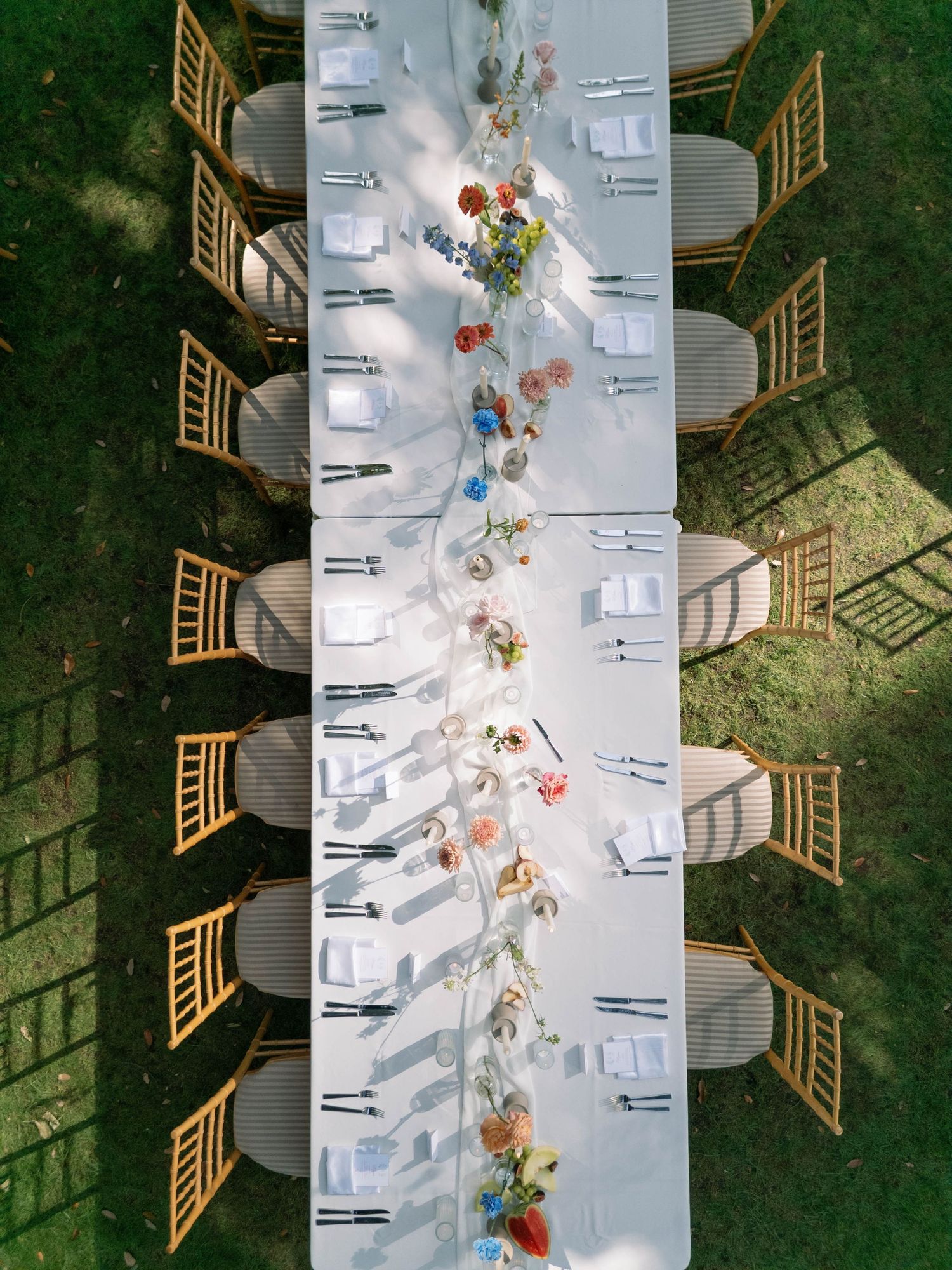 Aerial view of an elegant outdoor wedding table setting with white linens and wooden chairs on a green lawn.