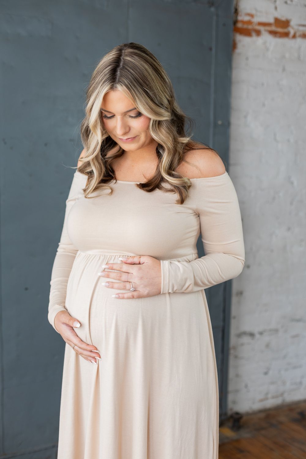 Maternity portrait of woman in off-shoulder cream dress against industrial backdrop.