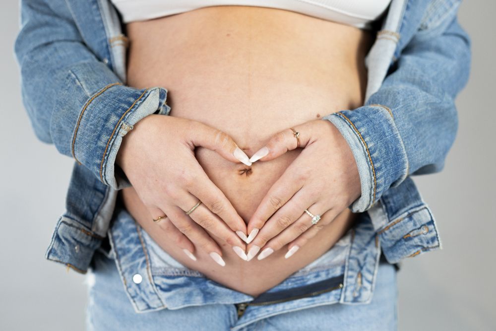 Hands forming heart shape over pregnant belly with denim jacket sleeves.