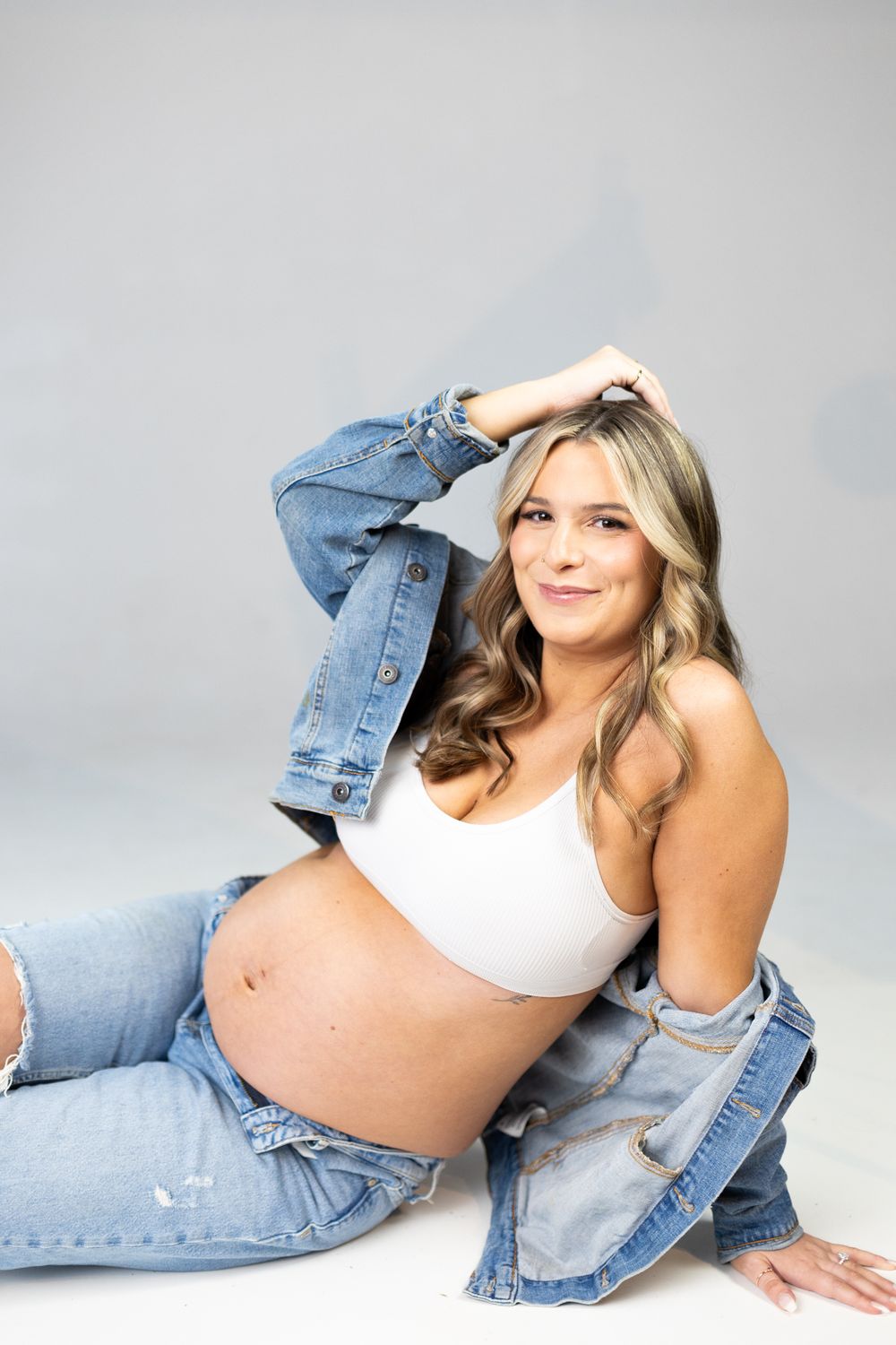 Maternity photo with denim jacket and white crop top against gray background.
