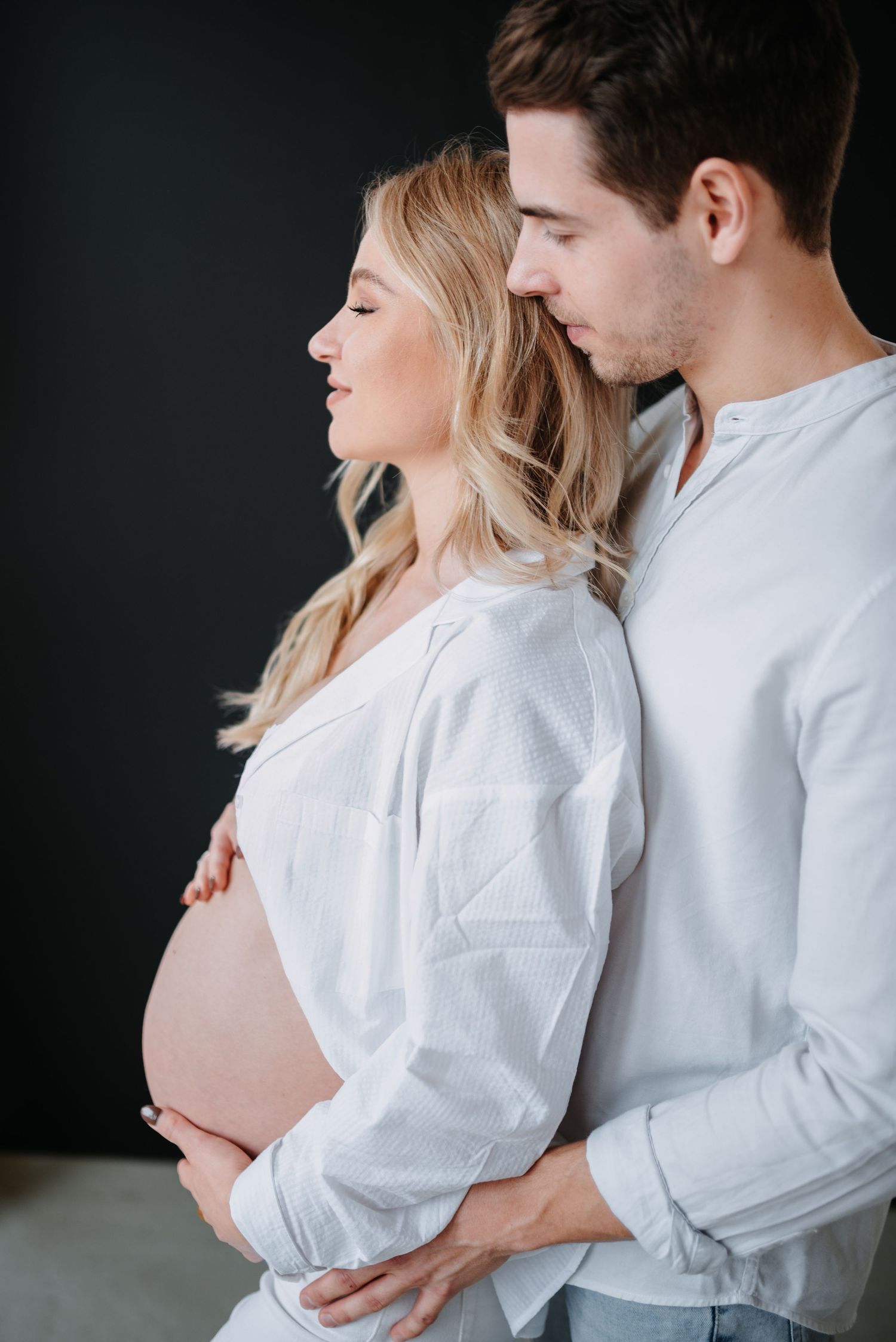 A romantic maternity photo of a couple in white shirts embracing against a dark background.