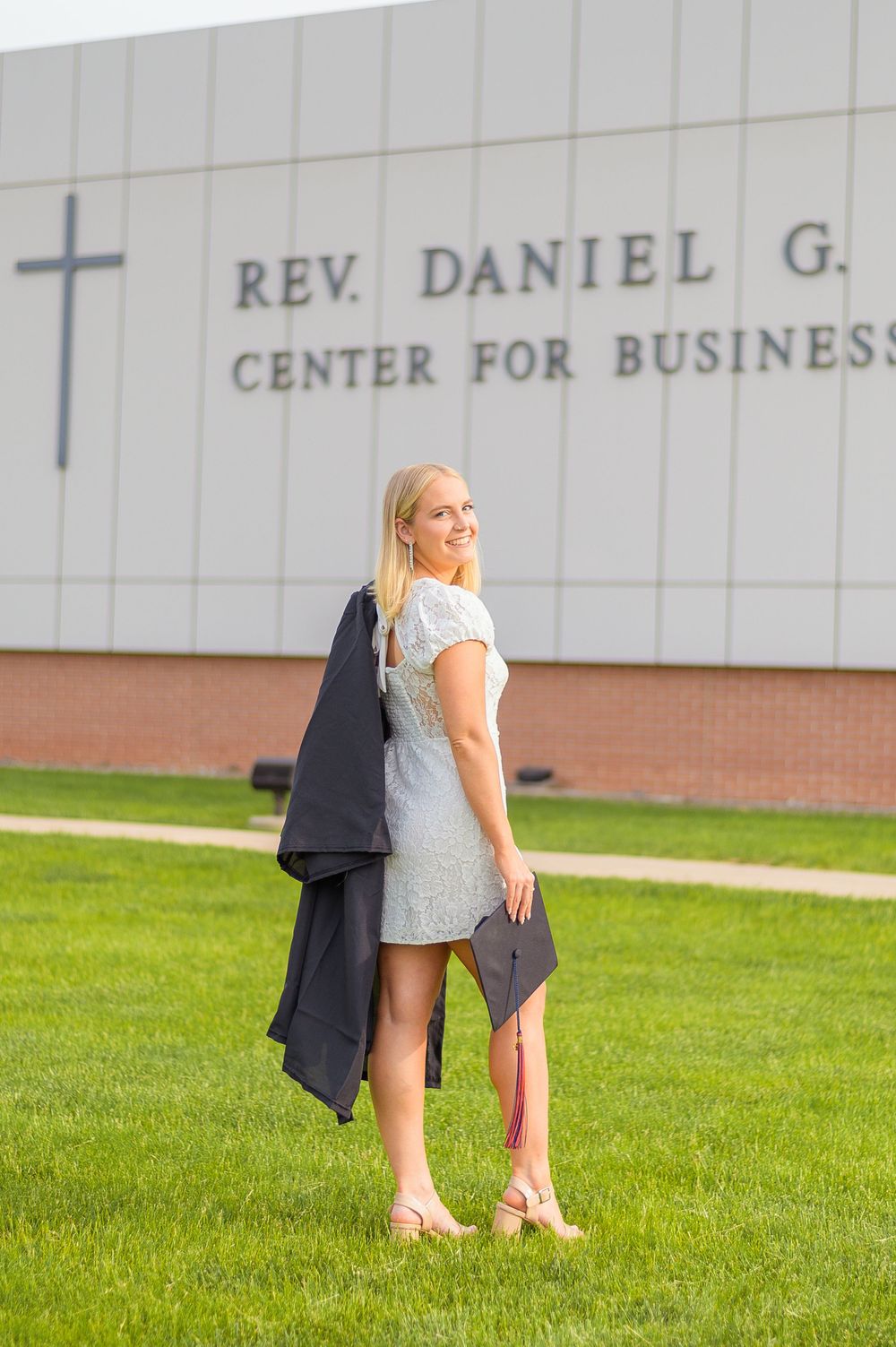 Graduate posing in a light dress outside the Rev. Daniel G. building at a university.
