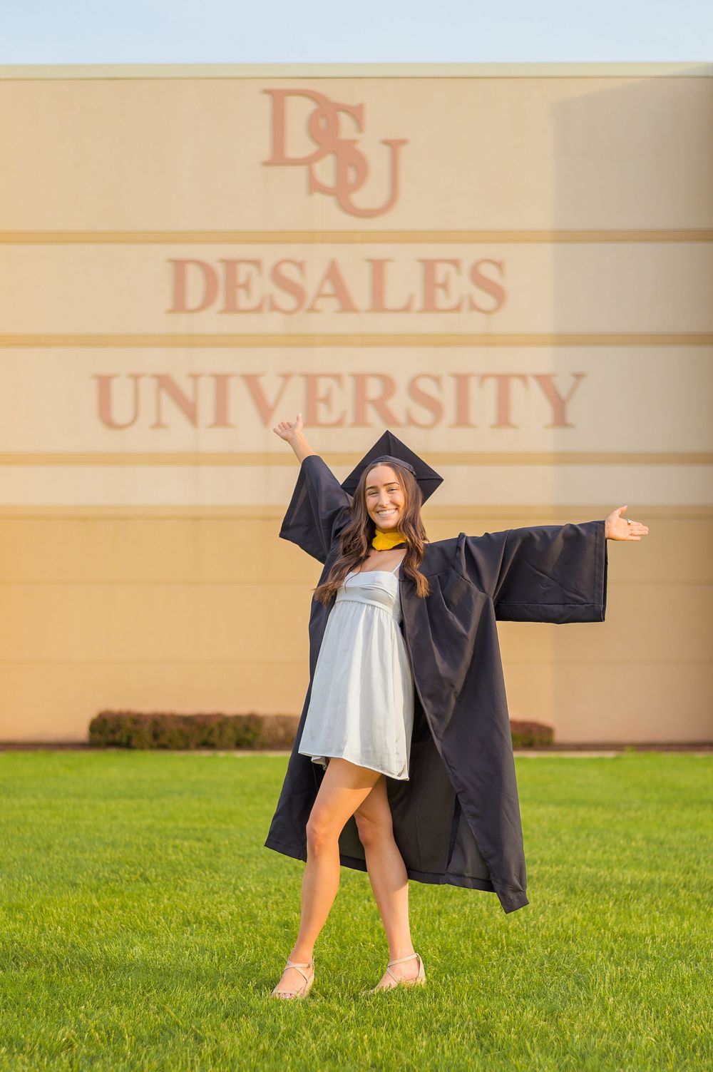 Graduate in cap and gown celebrating in front of DeSales University sign.