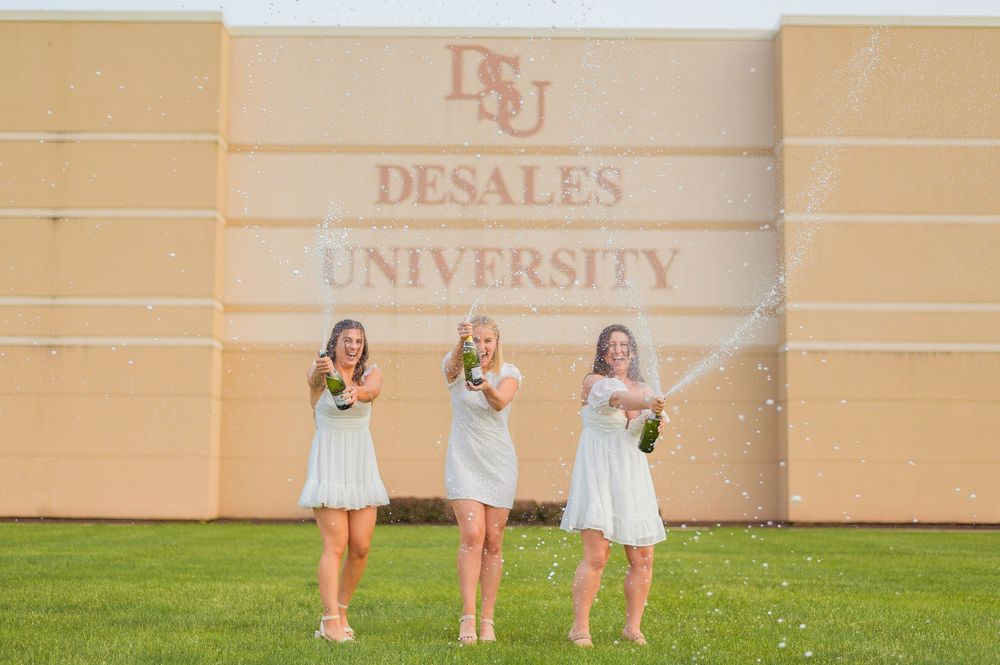 Three graduates in light dresses celebrate in front of DeSales University building.