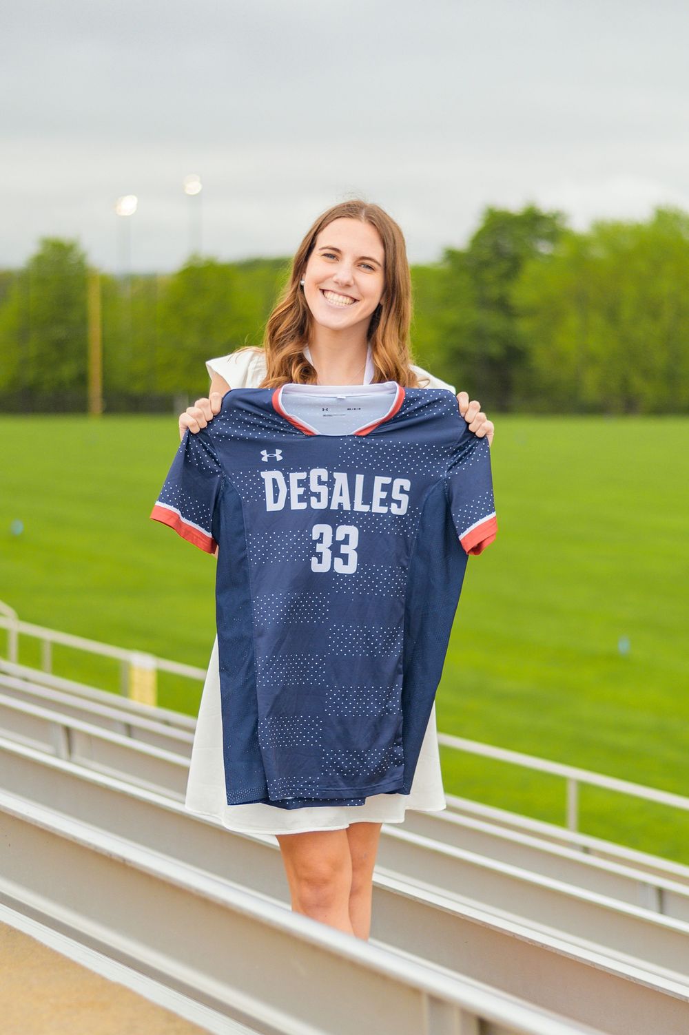 Graduate holding up blue DeSales athletic jersey number 33 on stadium bleachers.