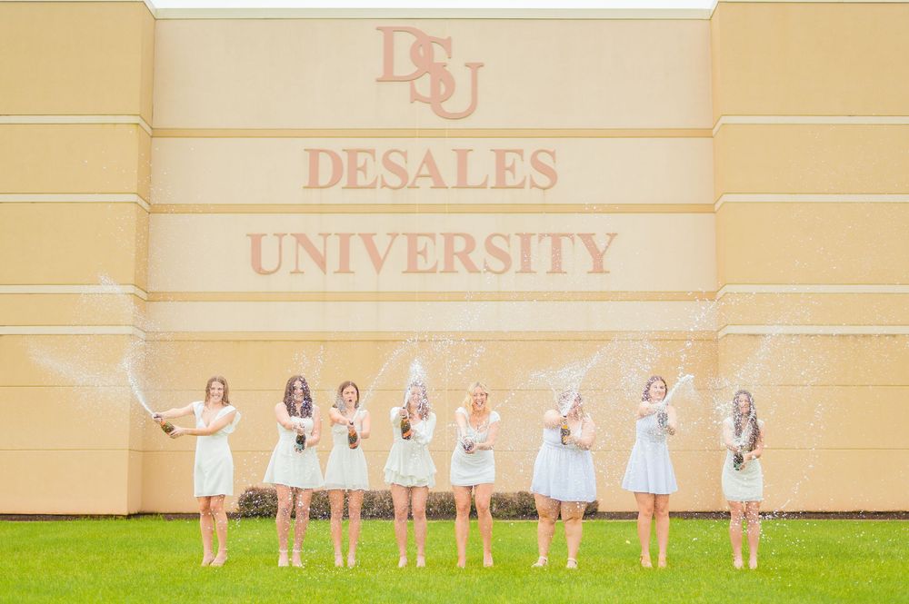 Graduates in light dresses pose in line against DeSales University building wall.