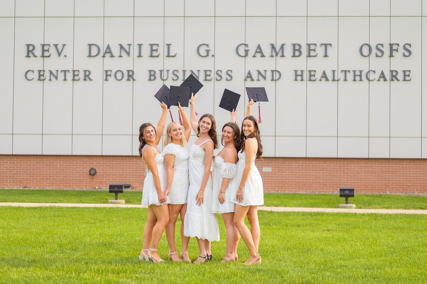 Graduates in white dresses toss their caps in celebration outside university building.