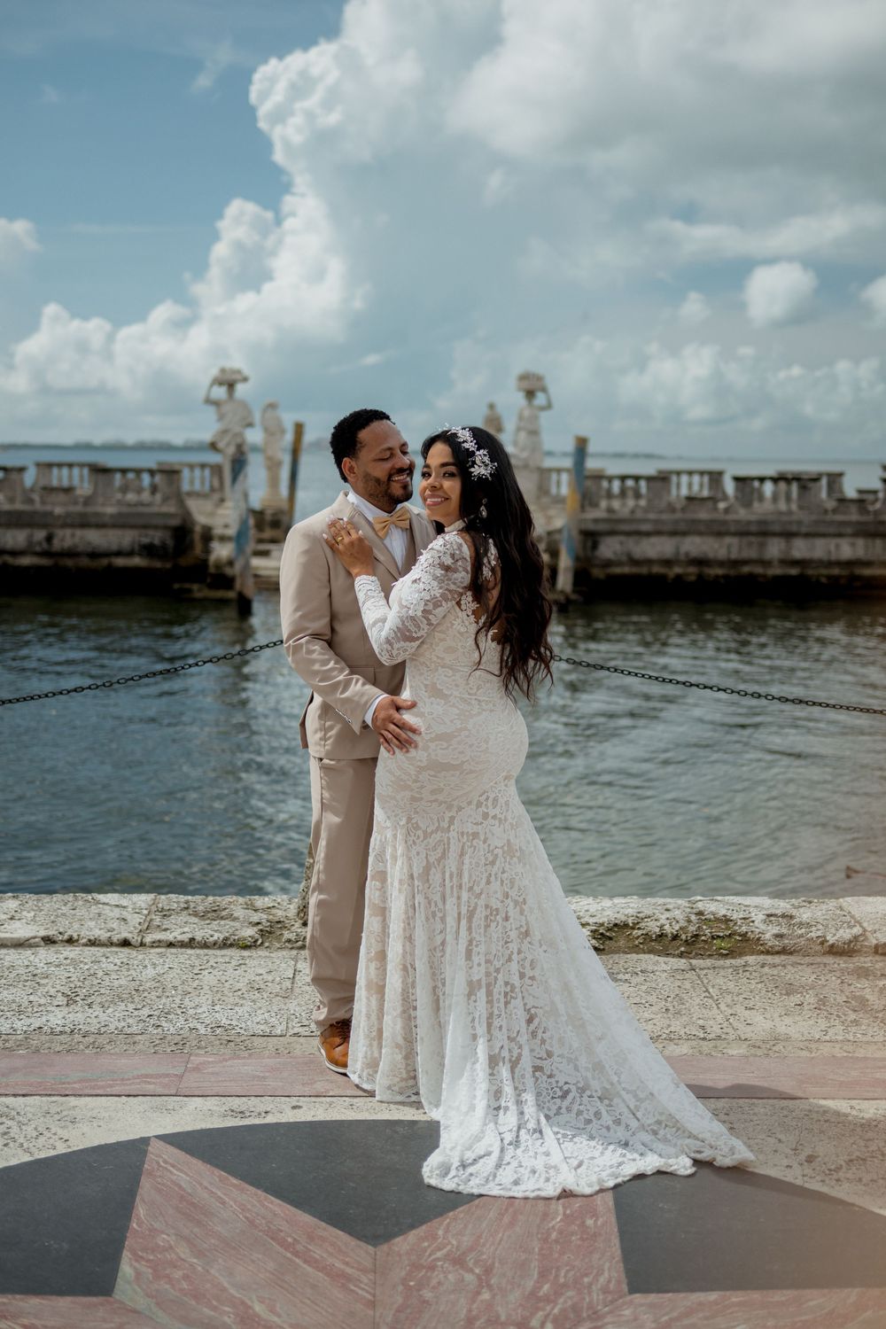 A couple in wedding attire embrace at a waterfront stone terrace with ornate pillars and cloudy blue skies in the background.