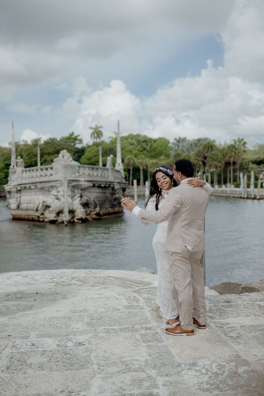 A couple in wedding attire embrace at the edge of a stone pool with an ornate stone boat fountain at Vizcaya Gardens.