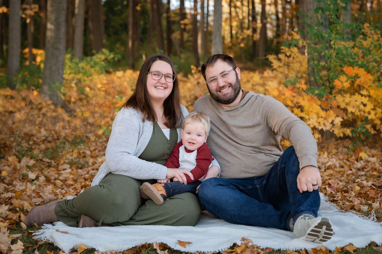 Family relaxing together on a blanket during an autumn photoshoot among fallen leaves in a sunlit forest setting.