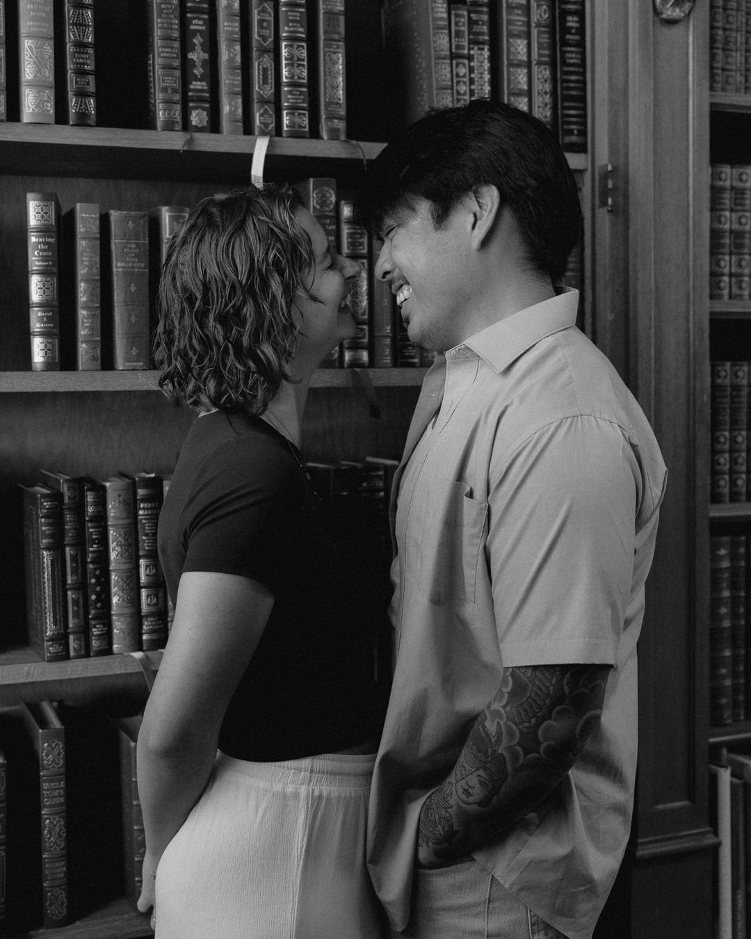 A couple that started laughing when they were about to kiss in front of a bookshelf.