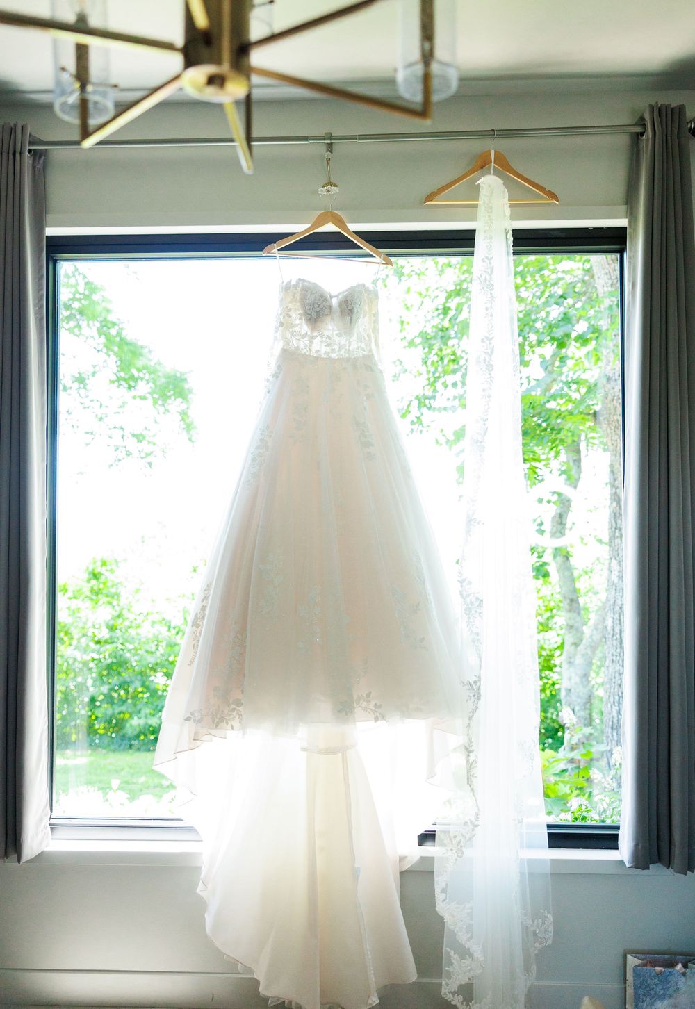 Elegant white wedding dress displayed hanging on wooden hangers against a window backdrop.