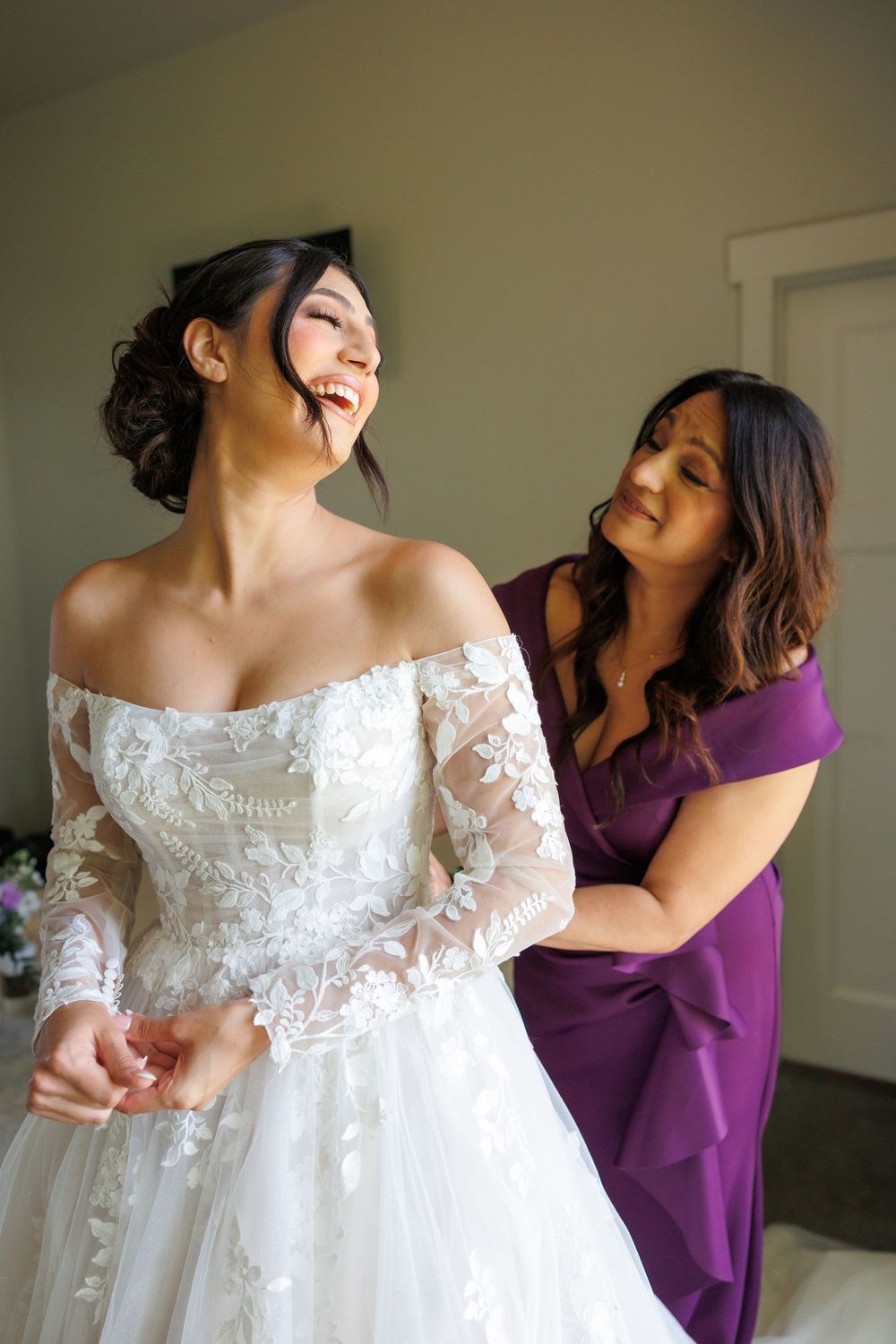 Wedding party helping bride into delicate lace off-shoulder wedding gown.