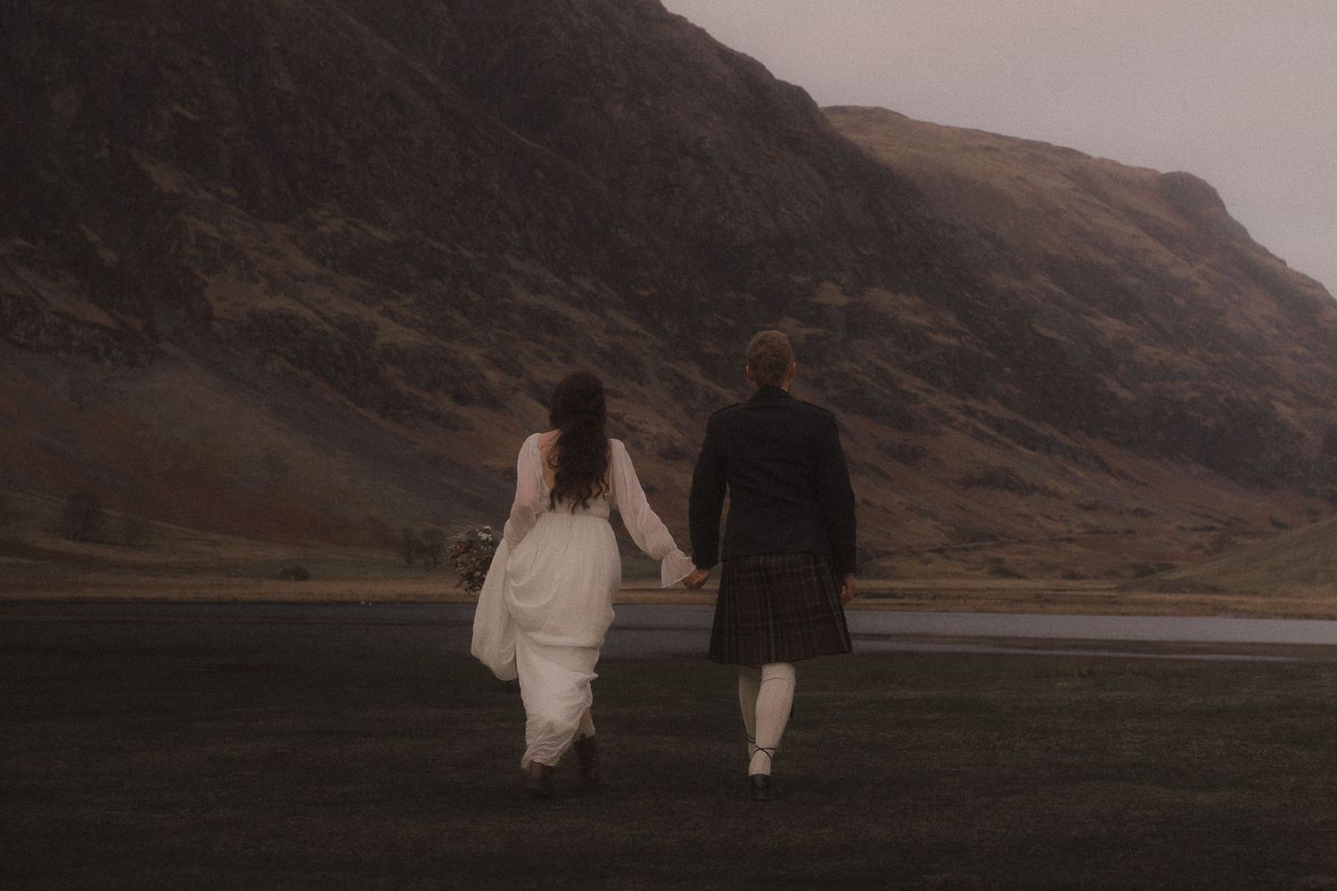 Natural moody photography of a couples at a misty lake beneath dramatic mountains in Glencoe Scotland during an elopement wedding
