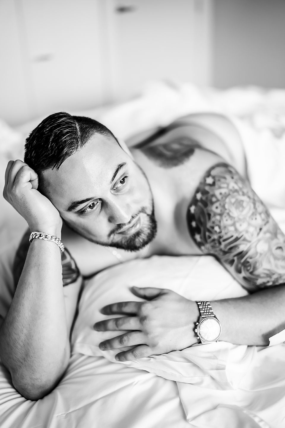 Black and white photo of person lying on white sheets with visible arm tattoos and wristwatch.