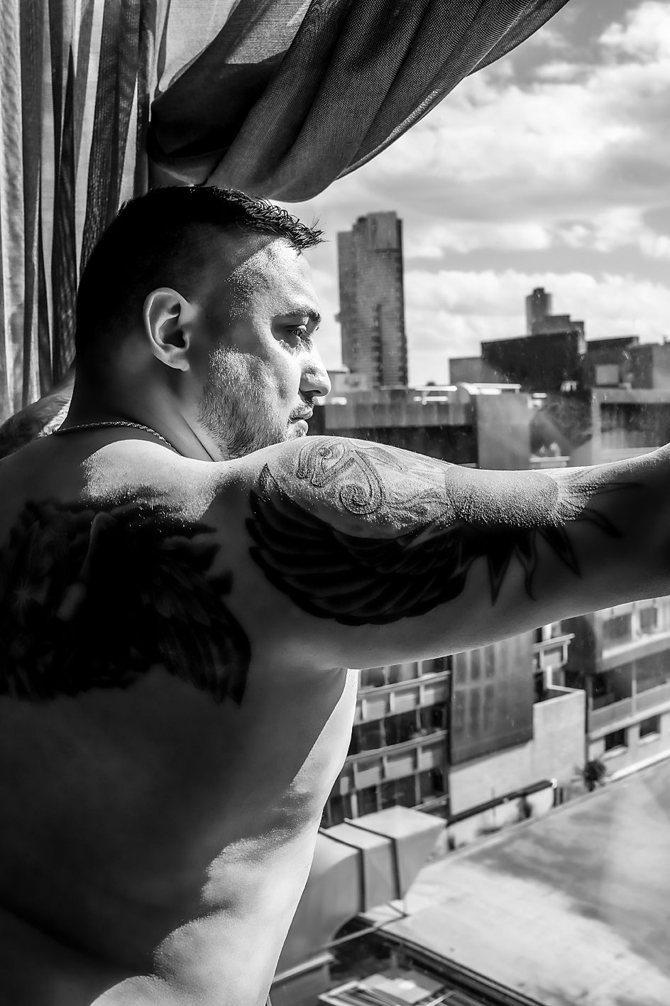 Black and white photo showing a tattooed arm resting on a window ledge overlooking a city with buildings in the distance.