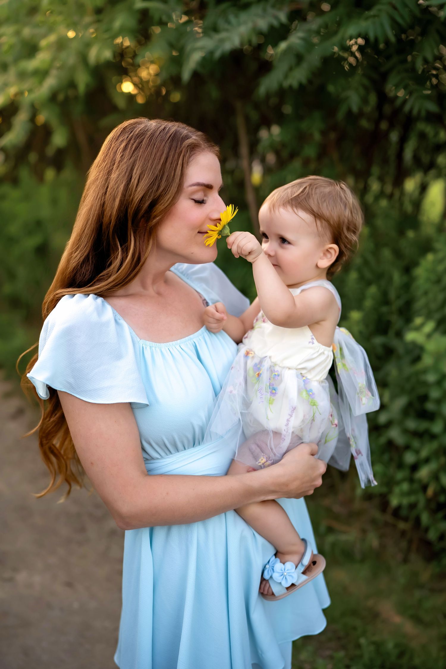 A woman in a light blue dress holds a baby in a floral outfit while sharing a tender moment outdoors in nature.
