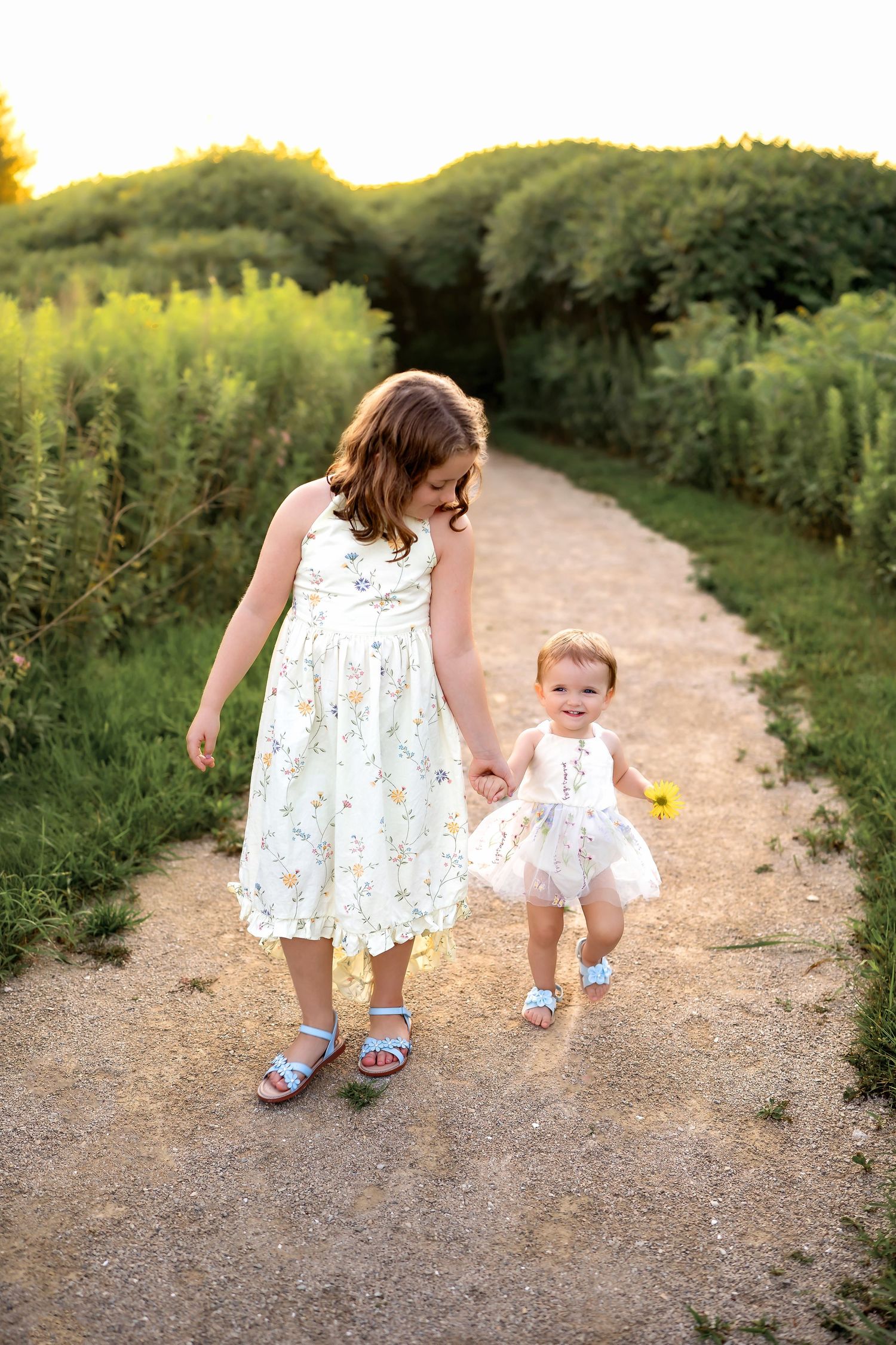 Two figures in matching floral dresses walk hand in hand down a nature path at sunset.