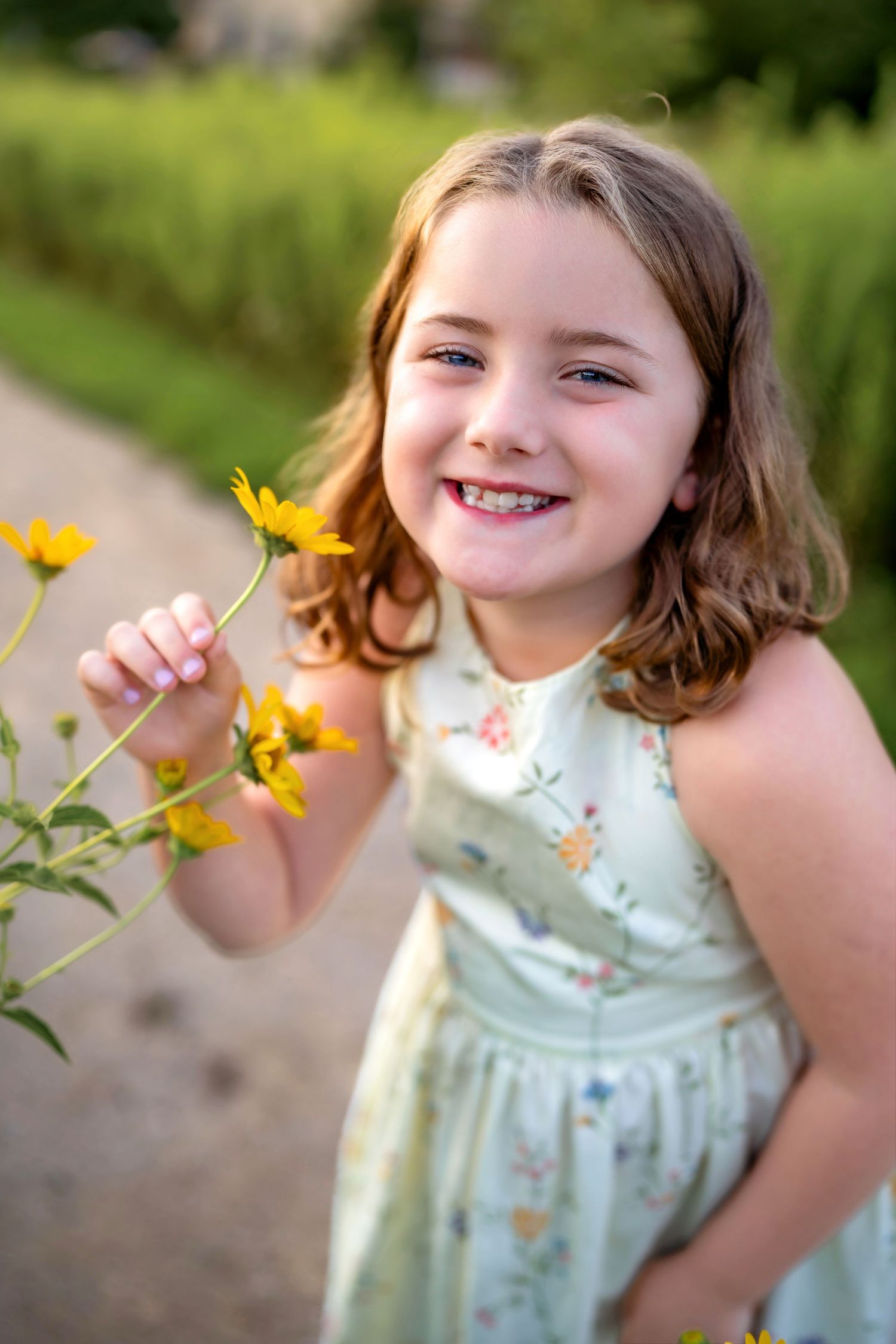 A young girl in a light-colored dress plays with yellow wildflowers in a sunny outdoor scene.