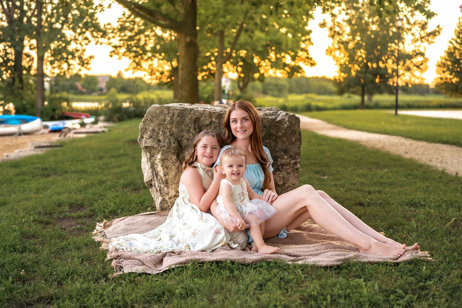 Three people sitting on a blanket outdoors during golden hour sunset at a park with trees in the background.