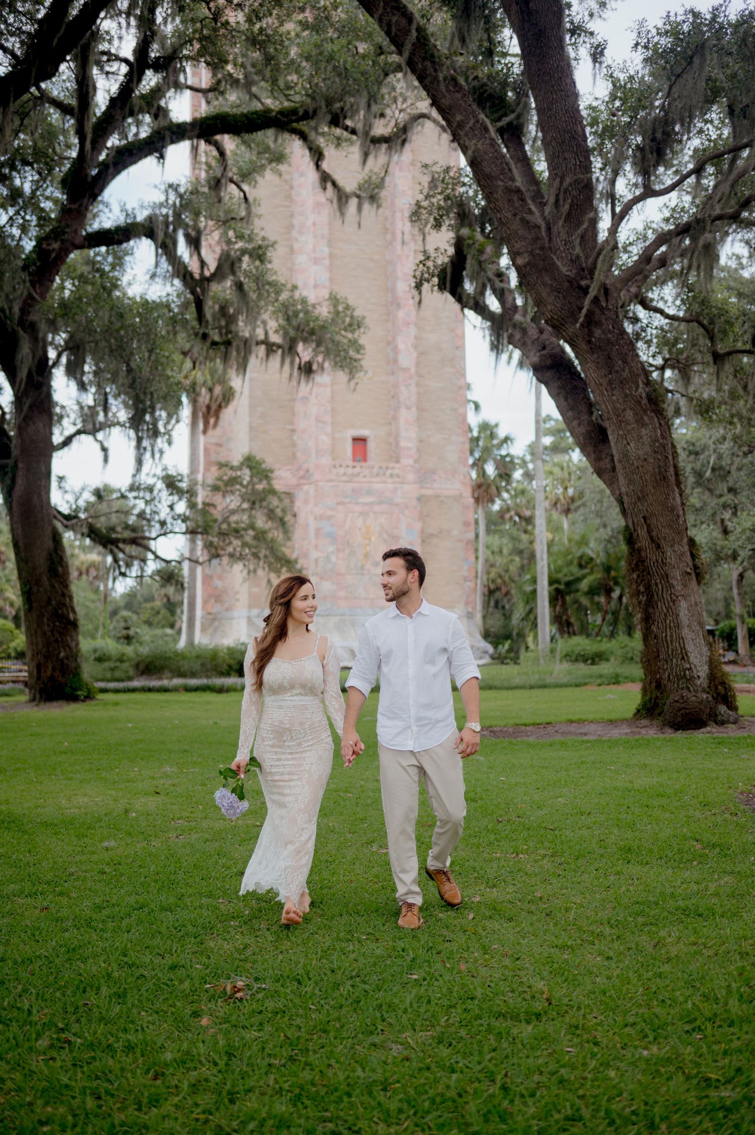 A couple strolls hand in hand on a lush green lawn with Spanish moss-draped oaks and a historic pink tower in the background.