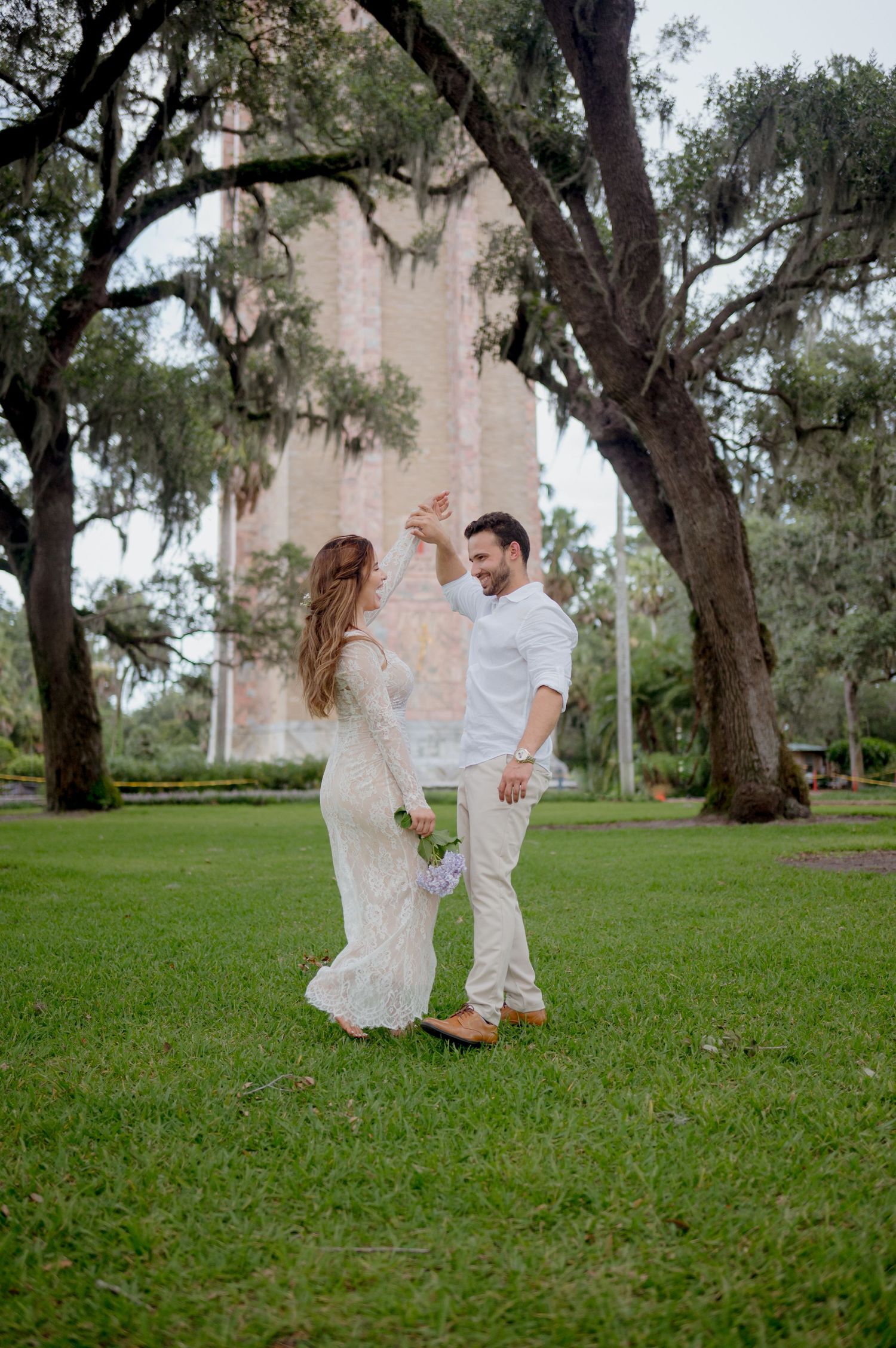 A couple in white outfits share a tender moment while dancing together on a grassy lawn under trees.