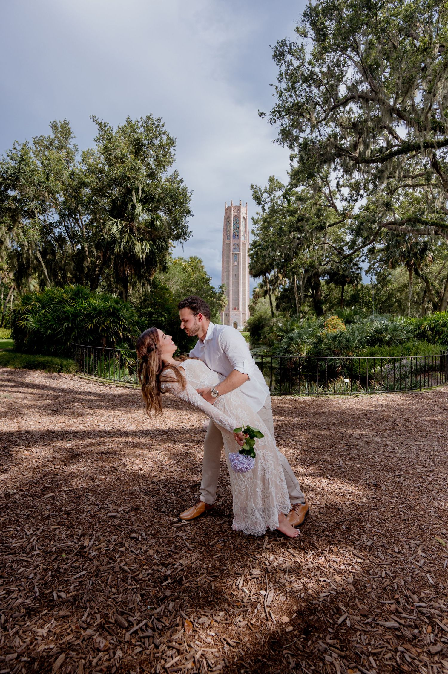 A romantic couple dips in their white wedding attire in front of Bok Tower Gardens surrounded by Spanish moss and oak trees.