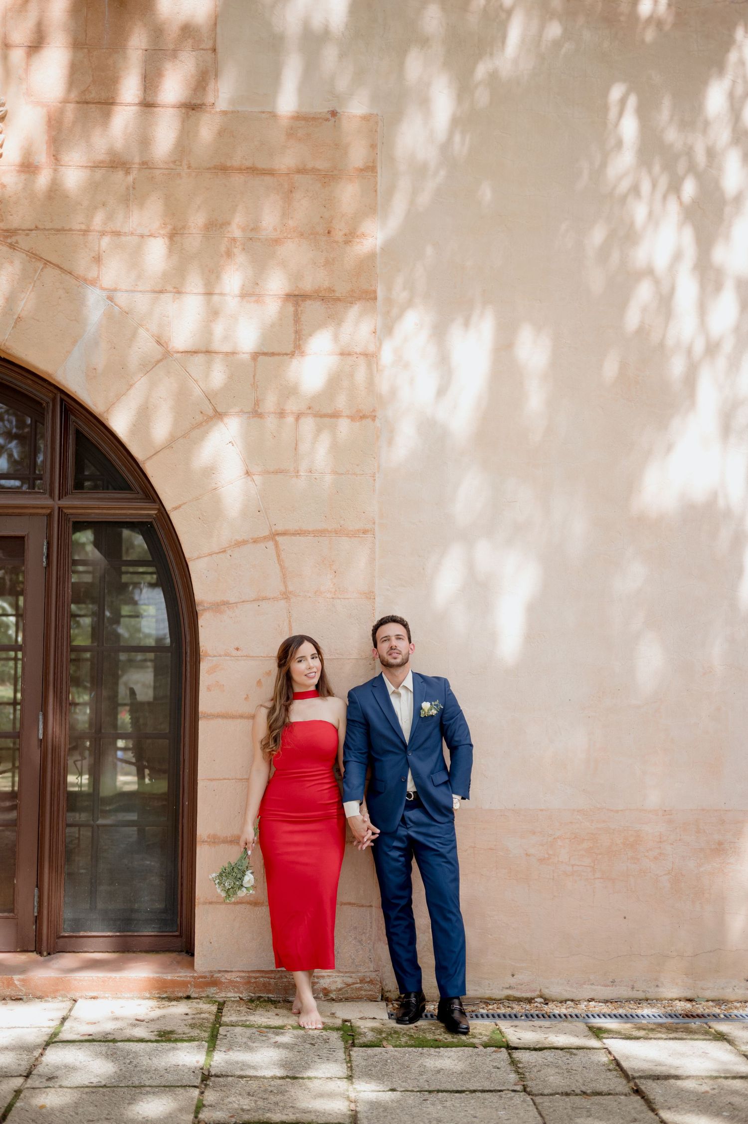 A couple in formal attire pose against a stone wall with dappled sunlight, wearing red and navy blue outfits.