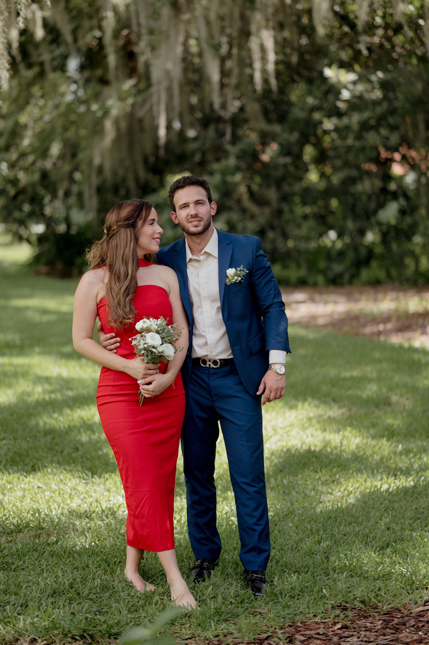 A romantic couple stand in a garden with Spanish moss, wearing a red dress and navy suit for a special occasion.