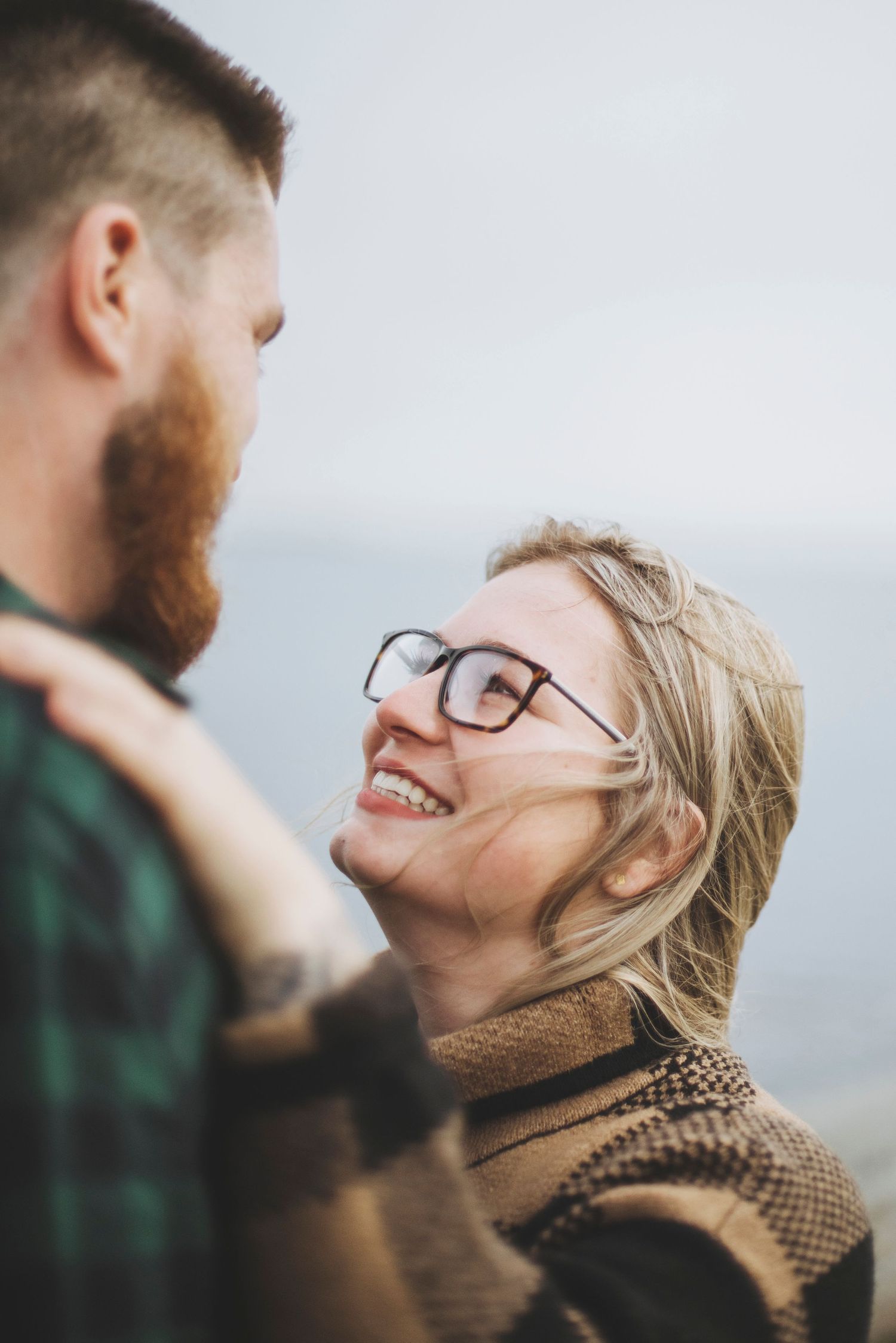 A woman looks up at her partner with a joyful smile, her hair gently tousled by the wind, as they share a warm moment.